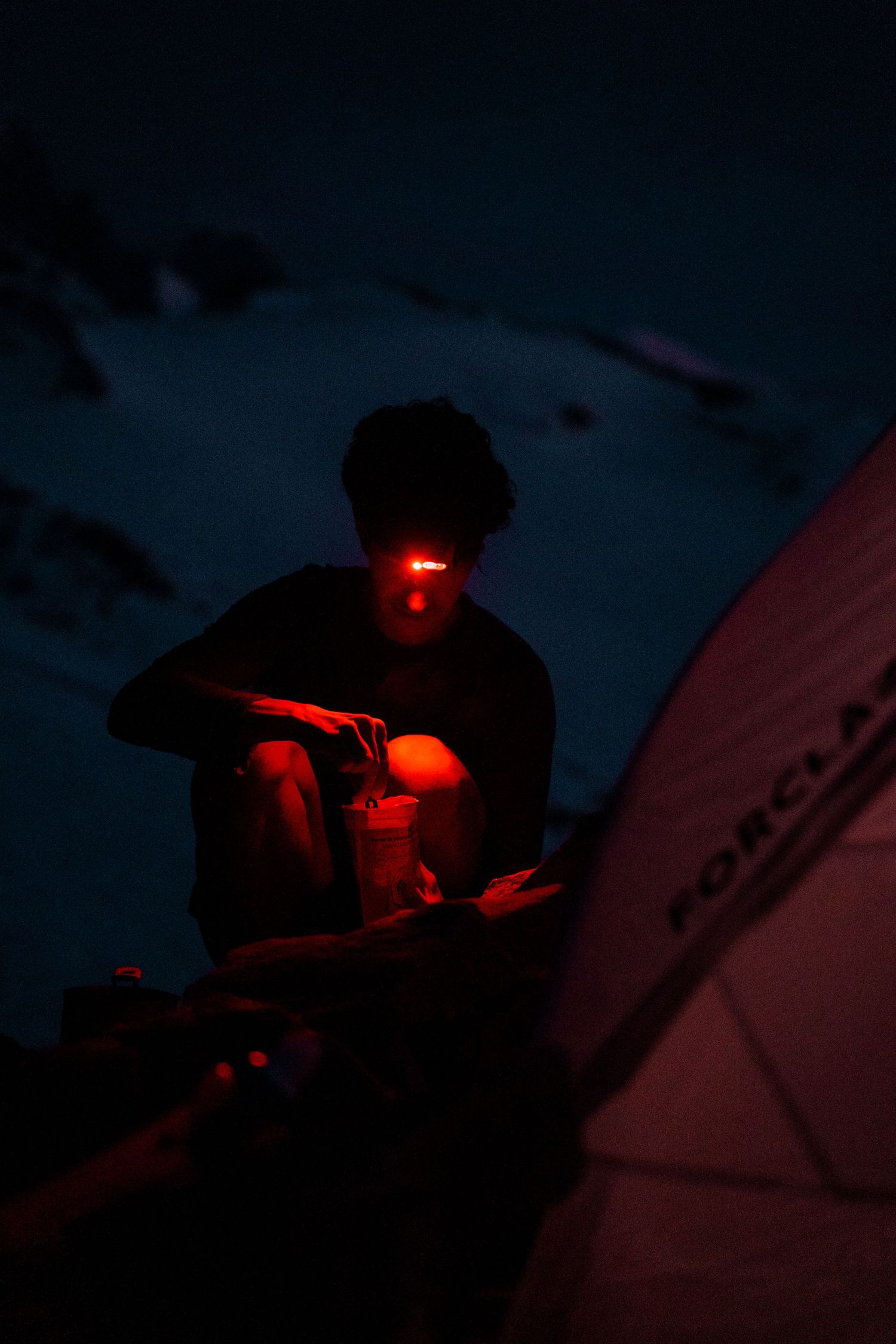 Mountain climber using red headlamp at night beside tent during high-altitude alpine camping expedition.