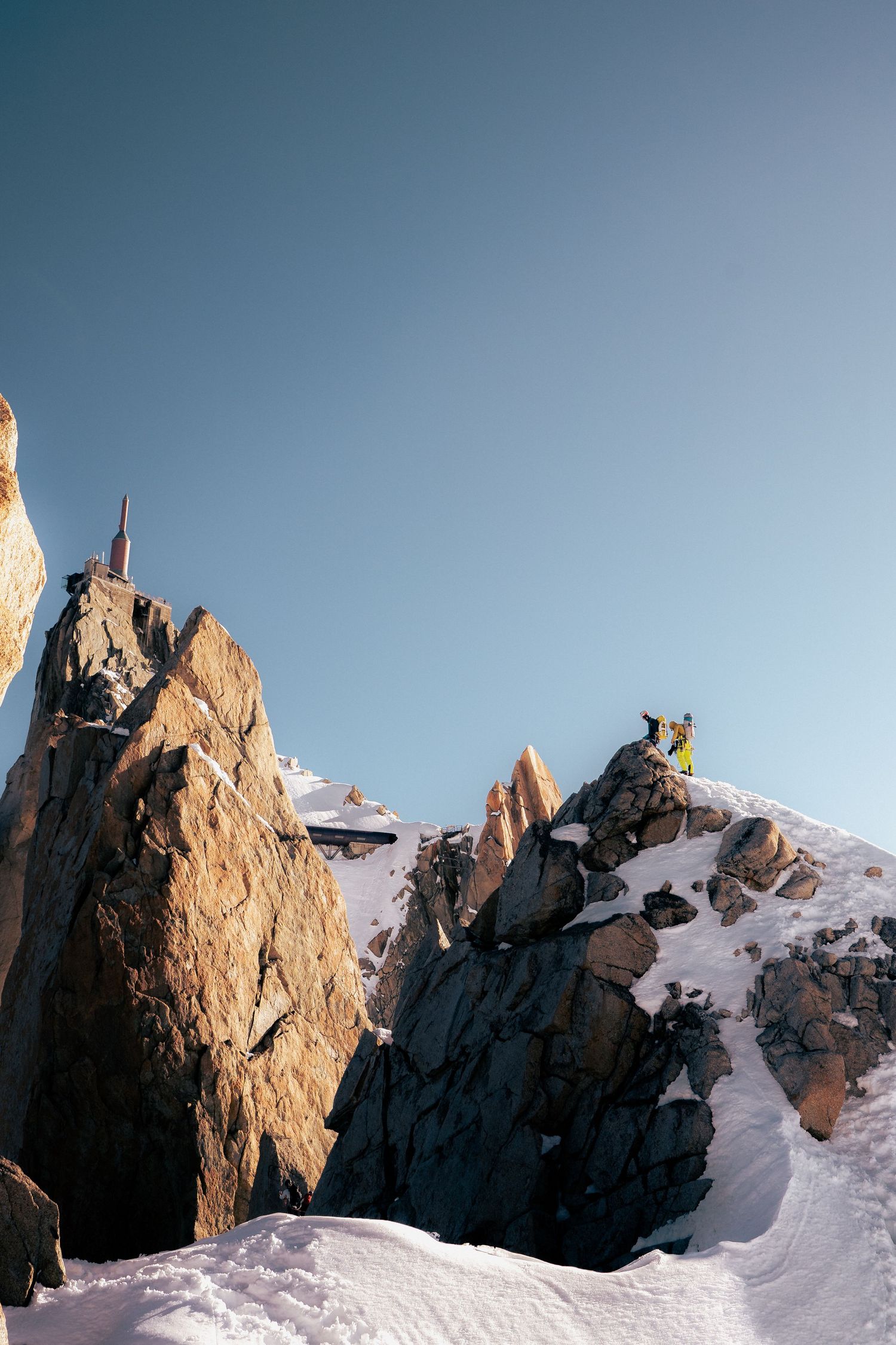 Climber in yellow jacket stands atop snow-covered rocky mountain peaks under clear blue sky.