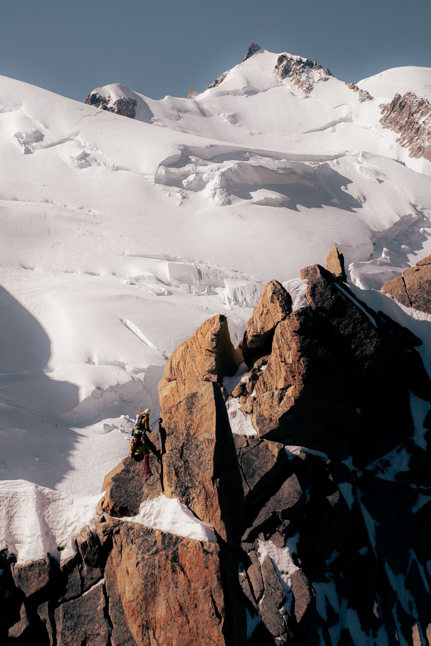 A mountaineer climbs jagged rocky peaks with a vast snow-covered glacier and mountain summit in the background.