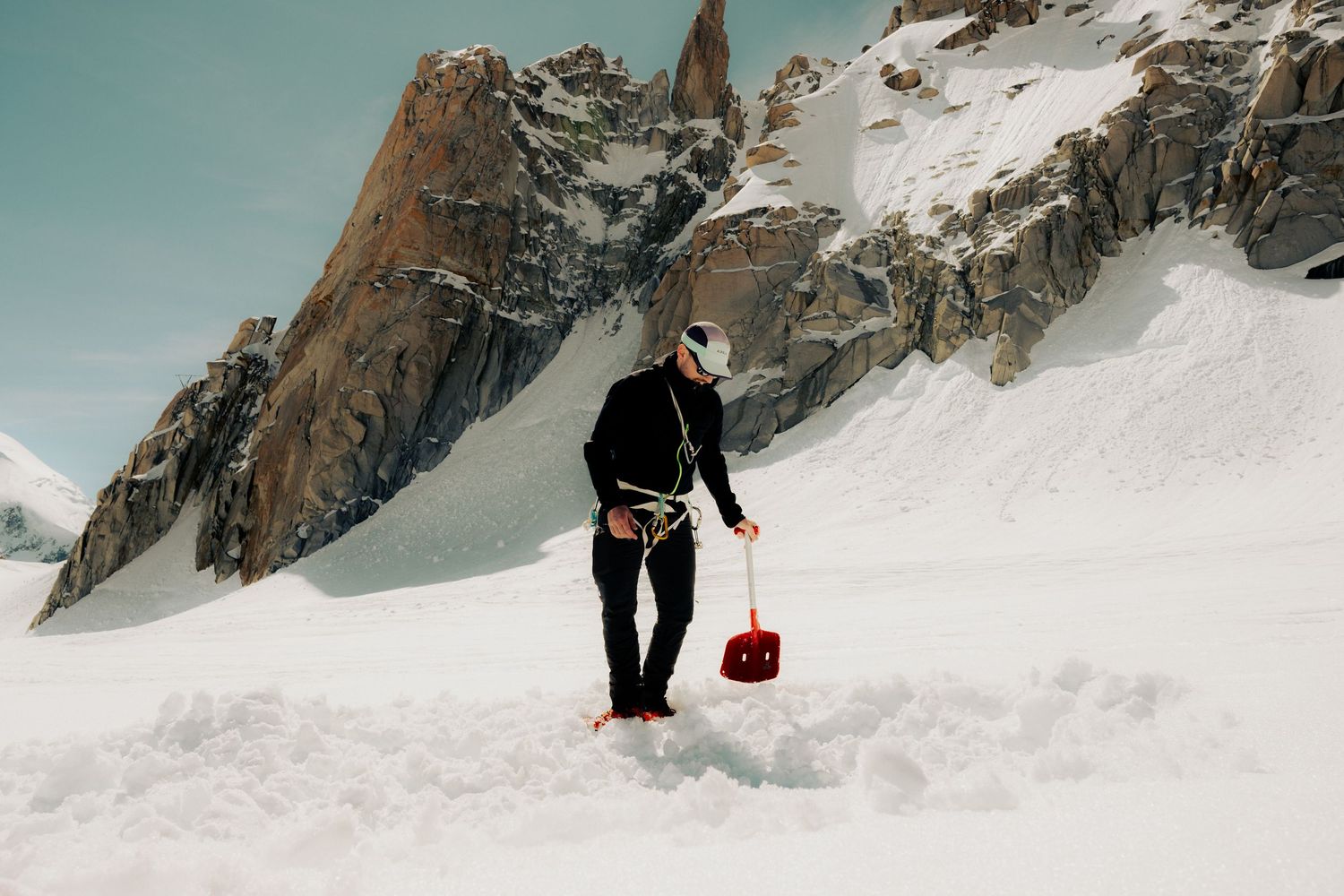 A mountaineer in black gear carries a red bag while trekking through deep snow with rocky peaks in the background.