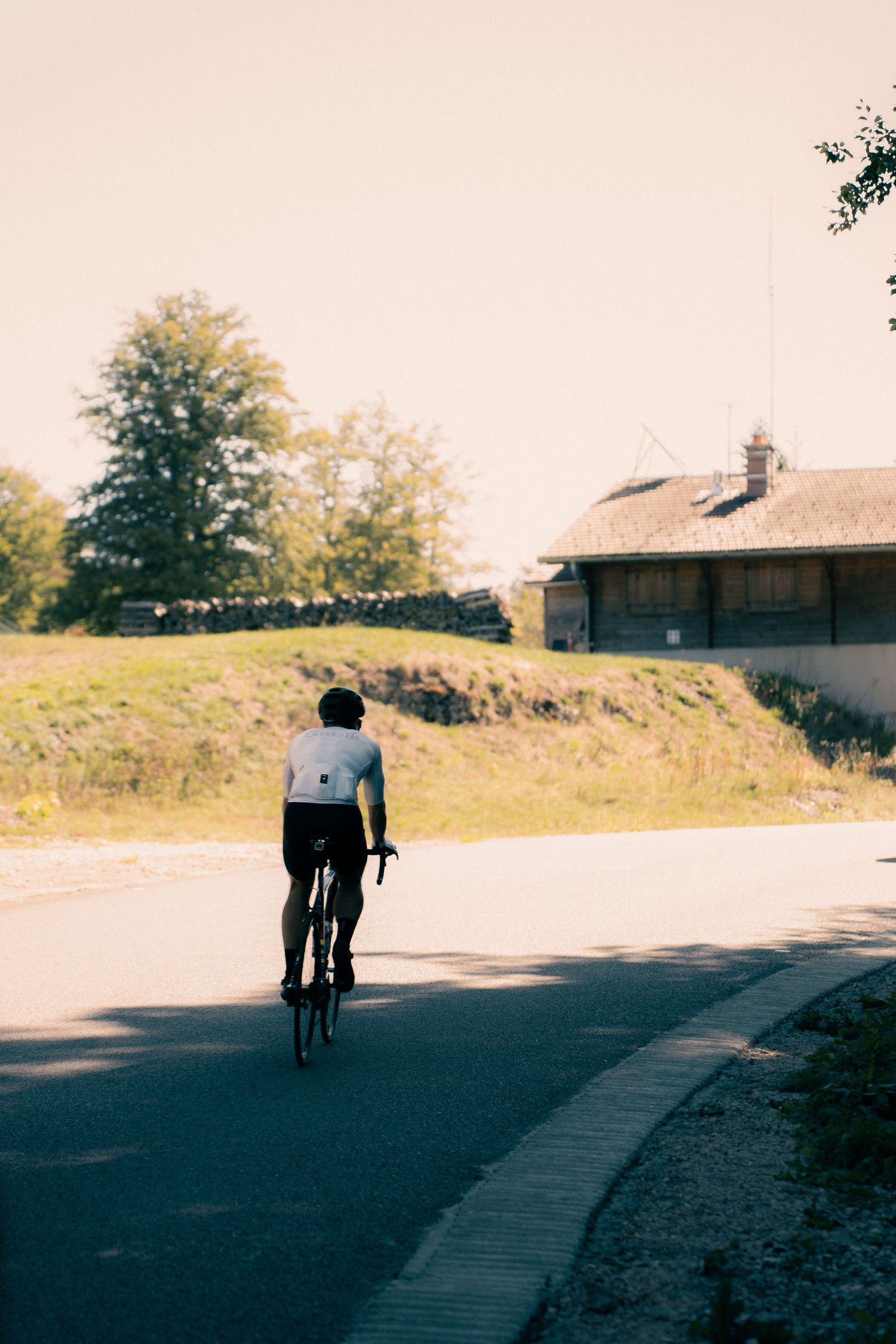 Cyclist riding a road bike away from camera on a quiet rural road with a stone building and trees in background.