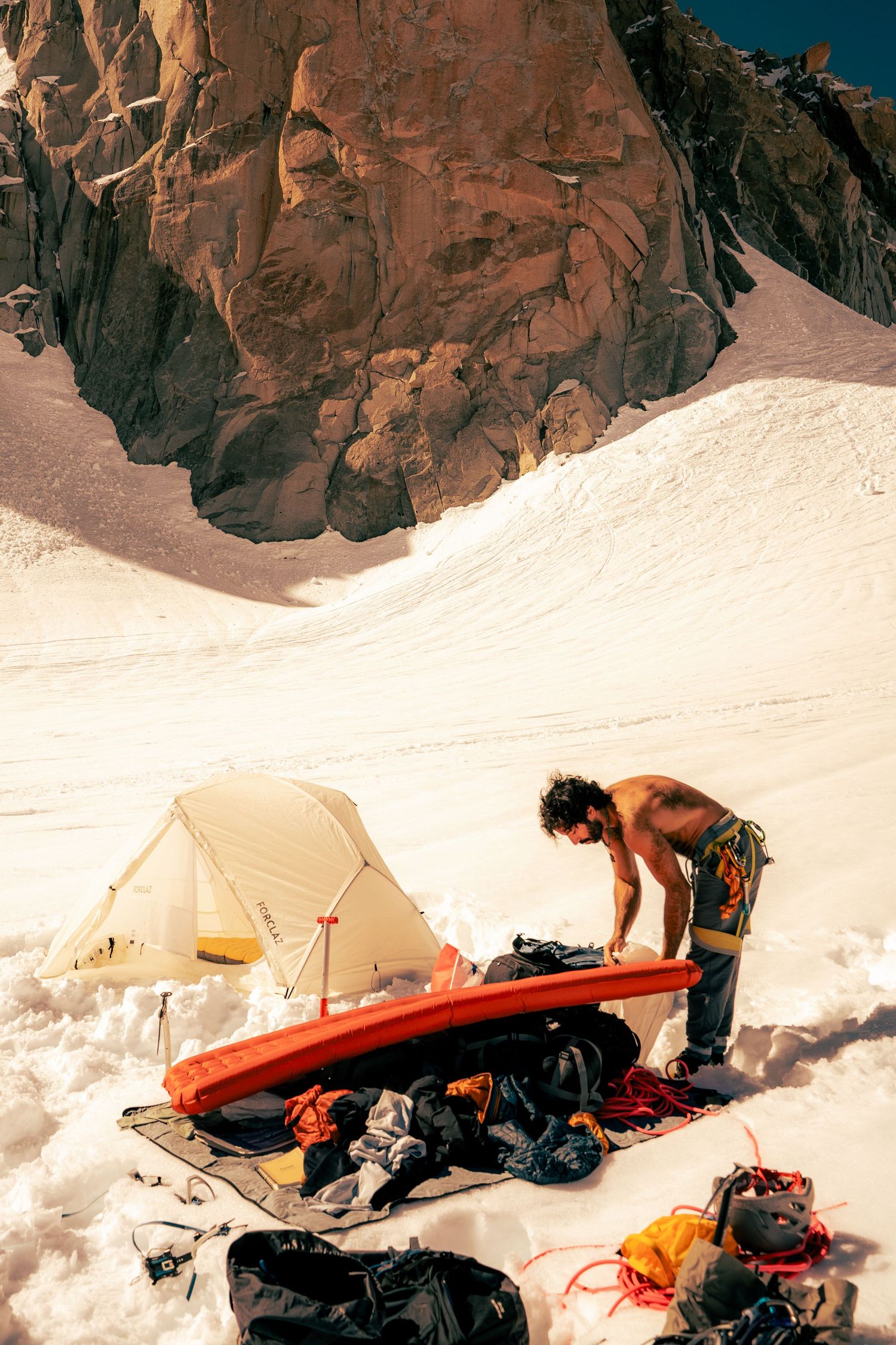 A shirtless climber organizes gear beside a tent on a snowy mountain slope with dramatic red rock cliffs behind.