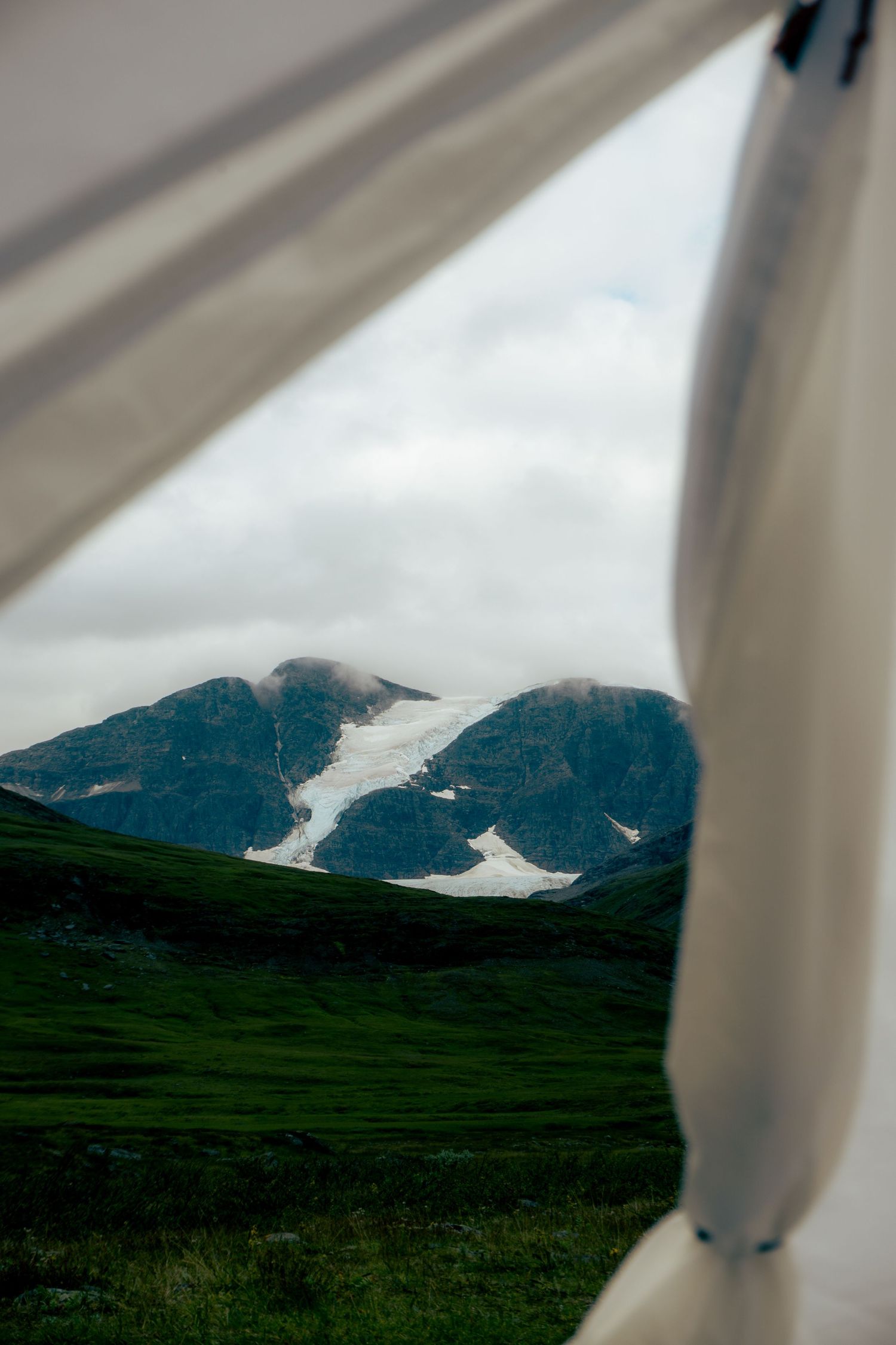 Snow-capped mountain peaks viewed through the open door of a white tent on a cloudy day.
