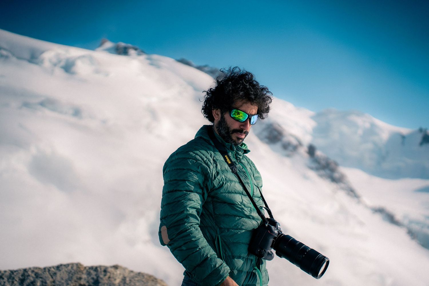 A bearded male photographer in a green jacket holds a camera on a snowy mountain summit under blue skies.