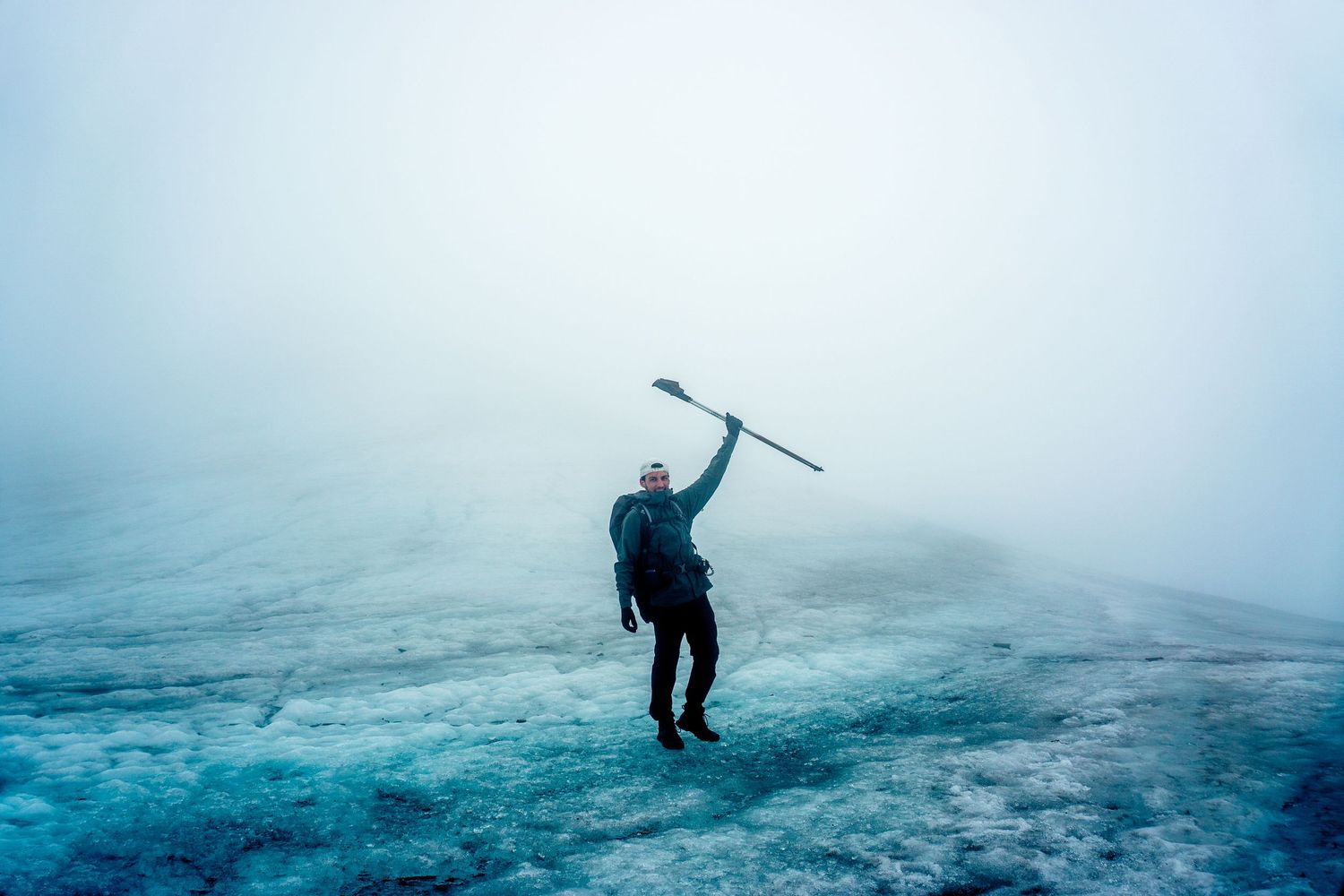 Hiker triumphantly raising an ice axe on a foggy glacier in remote arctic wilderness.
