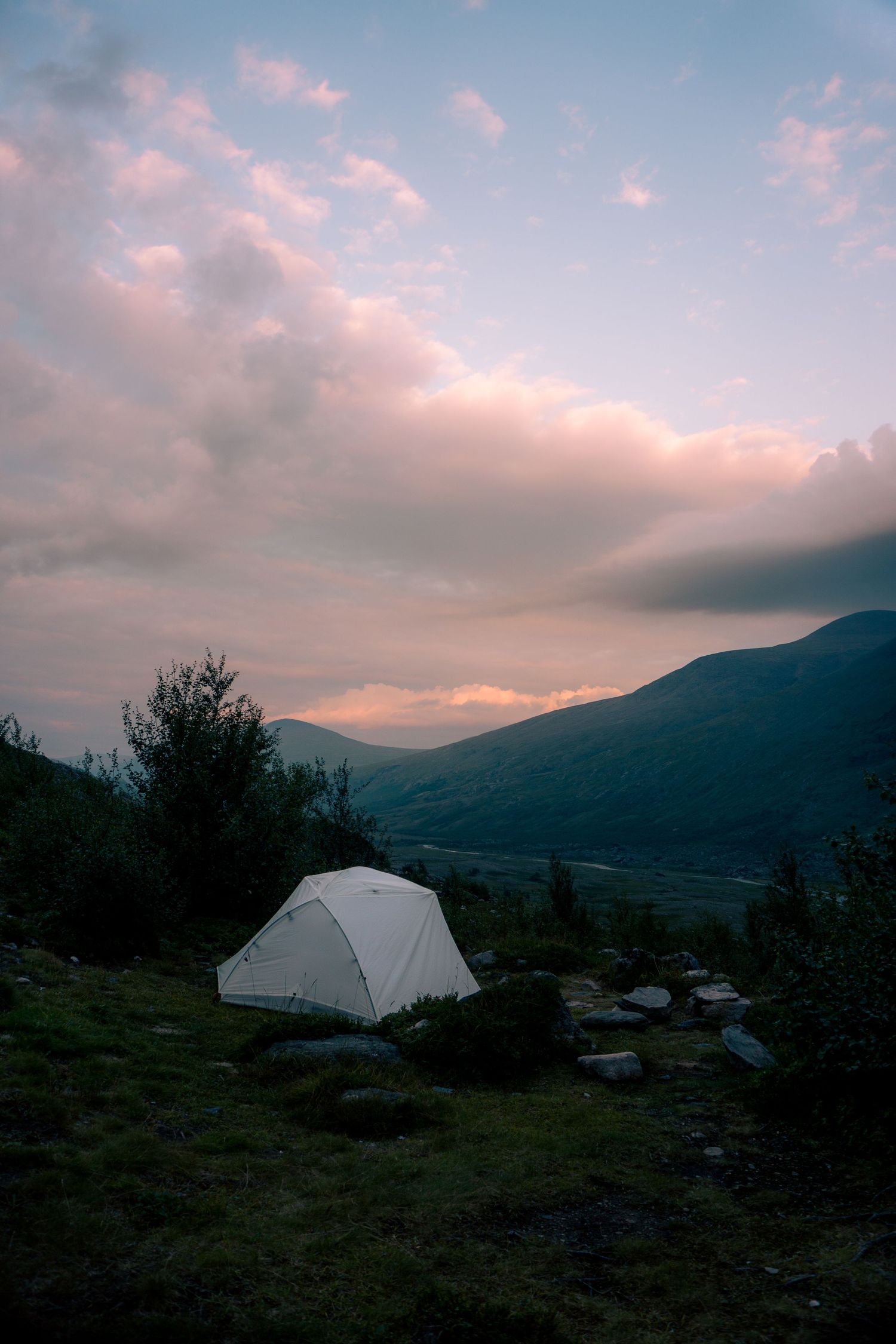 White camping tent pitched on a hillside at sunset with mountain valley views and colorful clouds.