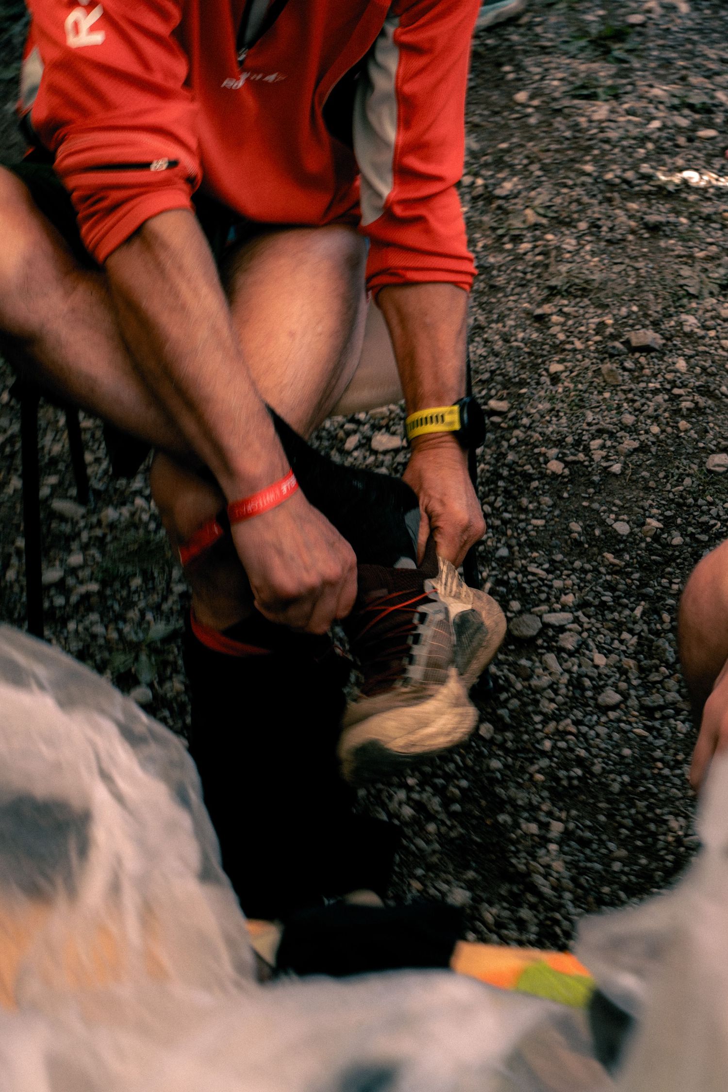 Trail runner in red jacket tying running shoe laces at a nighttime ultramarathon aid station.