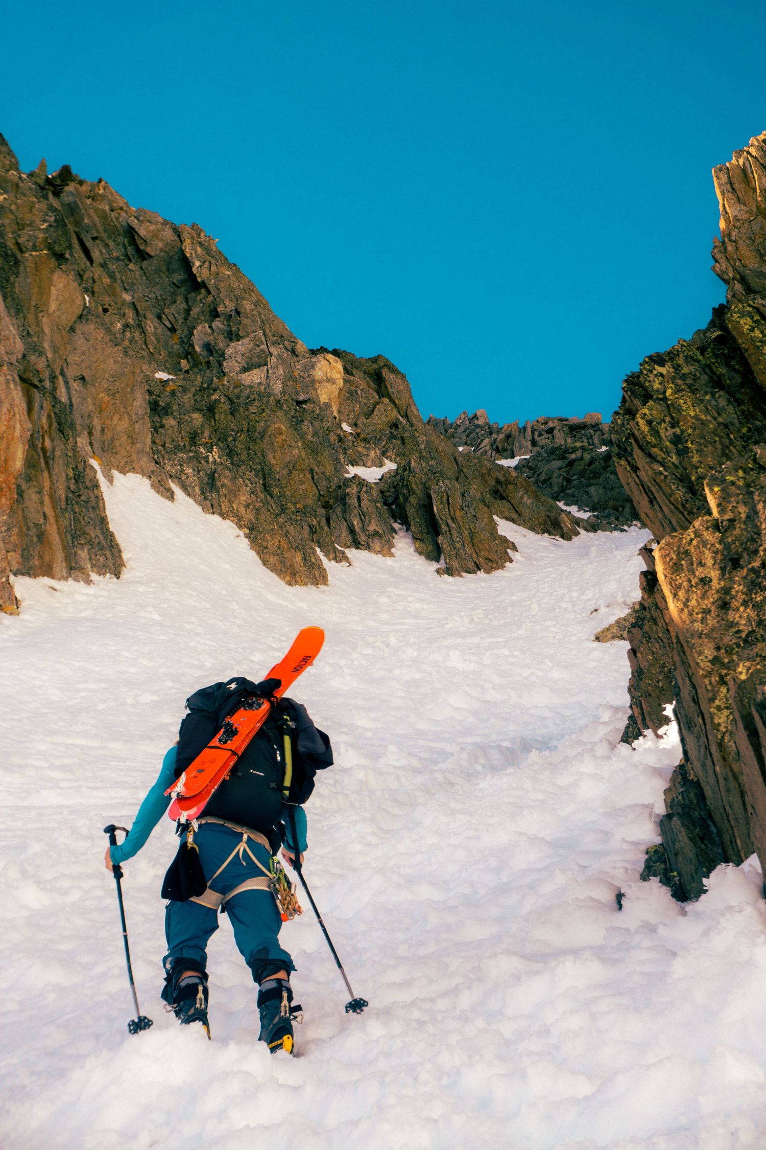 A mountaineer with skis on backpack climbs a steep snow couloir between rocky cliffs under a clear blue sky.