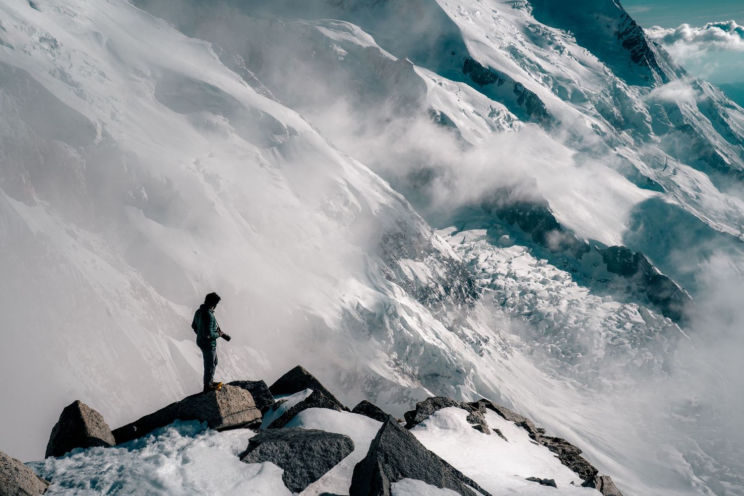 A lone figure stands on a rocky ridge overlooking vast snow-covered Alpine peaks wrapped in swirling clouds.