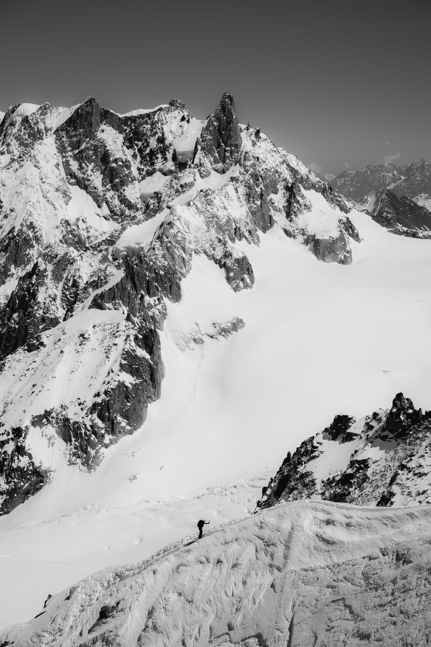Black and white photo of a lone mountaineer ascending a vast snowy alpine glacier with dramatic rocky peaks.