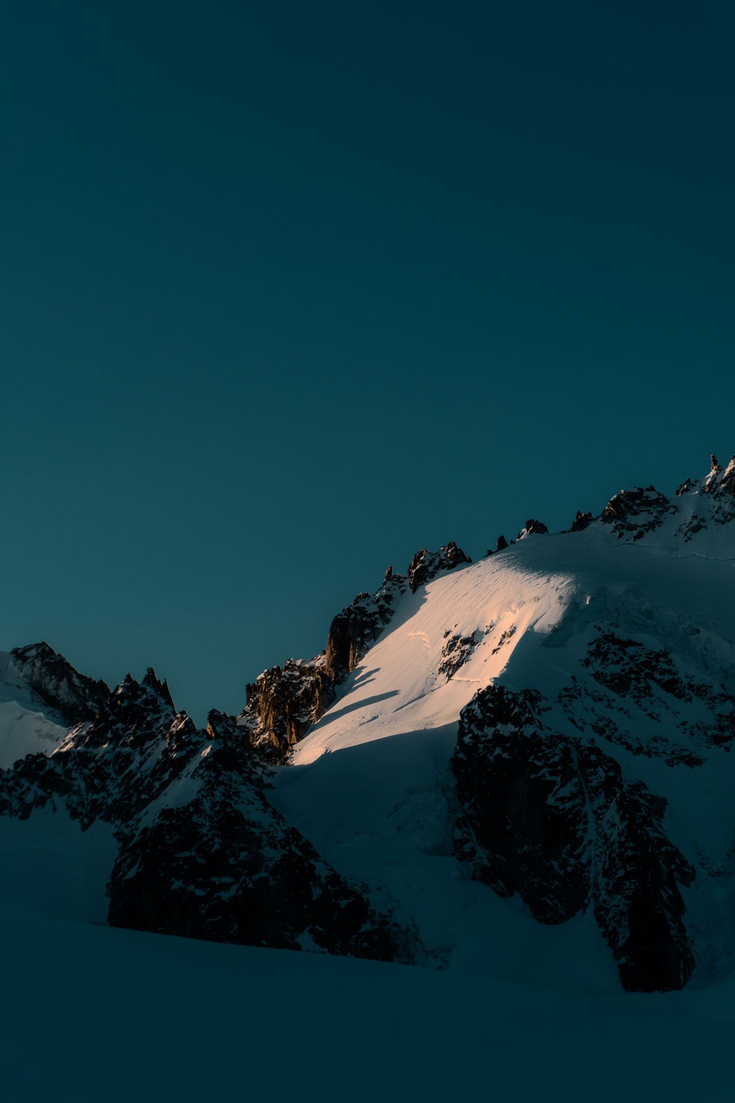 Snow-covered mountain peaks bathed in golden alpenglow light against a deep teal twilight sky.