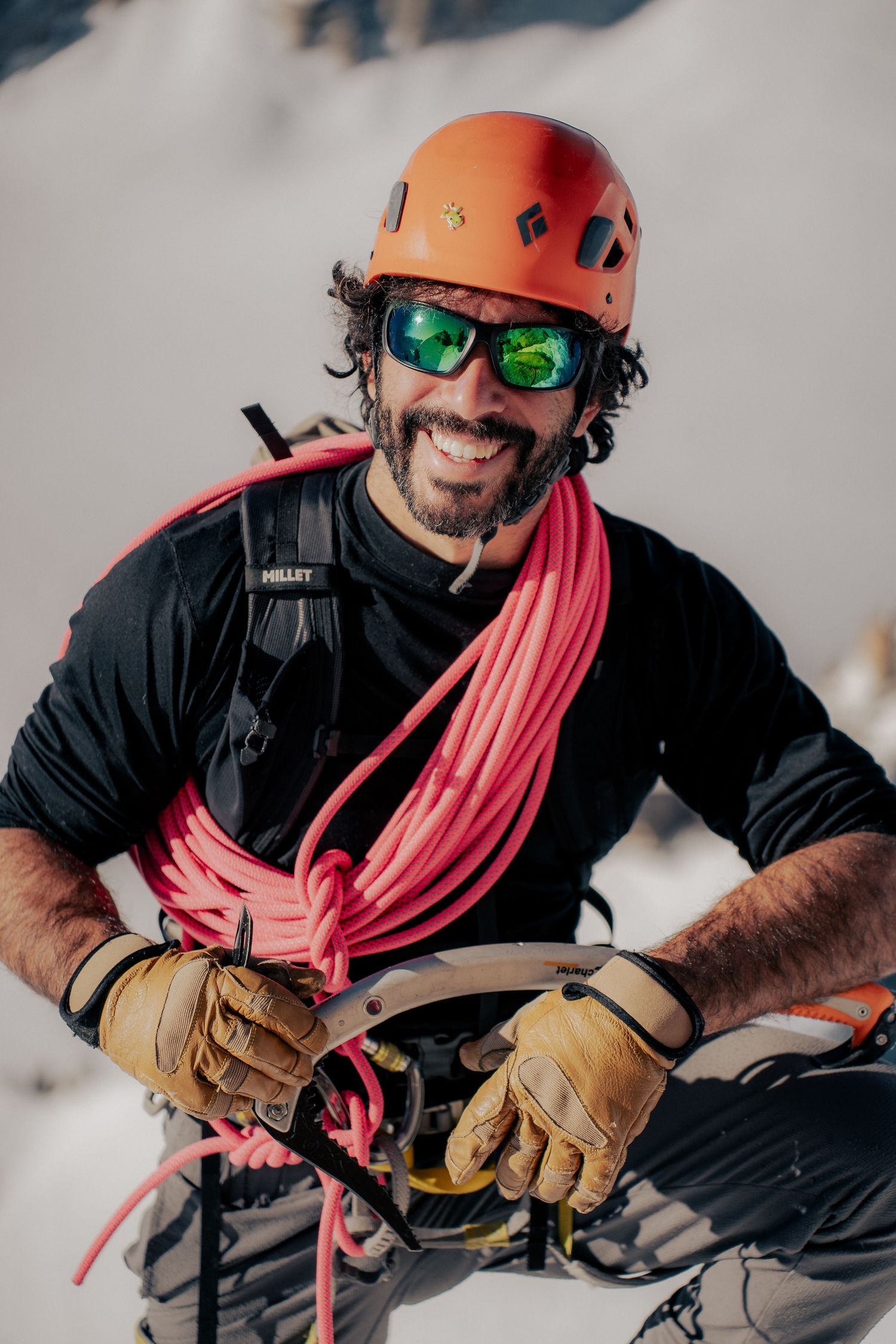 Smiling male rock climber wearing orange helmet, green mirrored sunglasses, and pink rope over shoulder in snowy mountain setting.