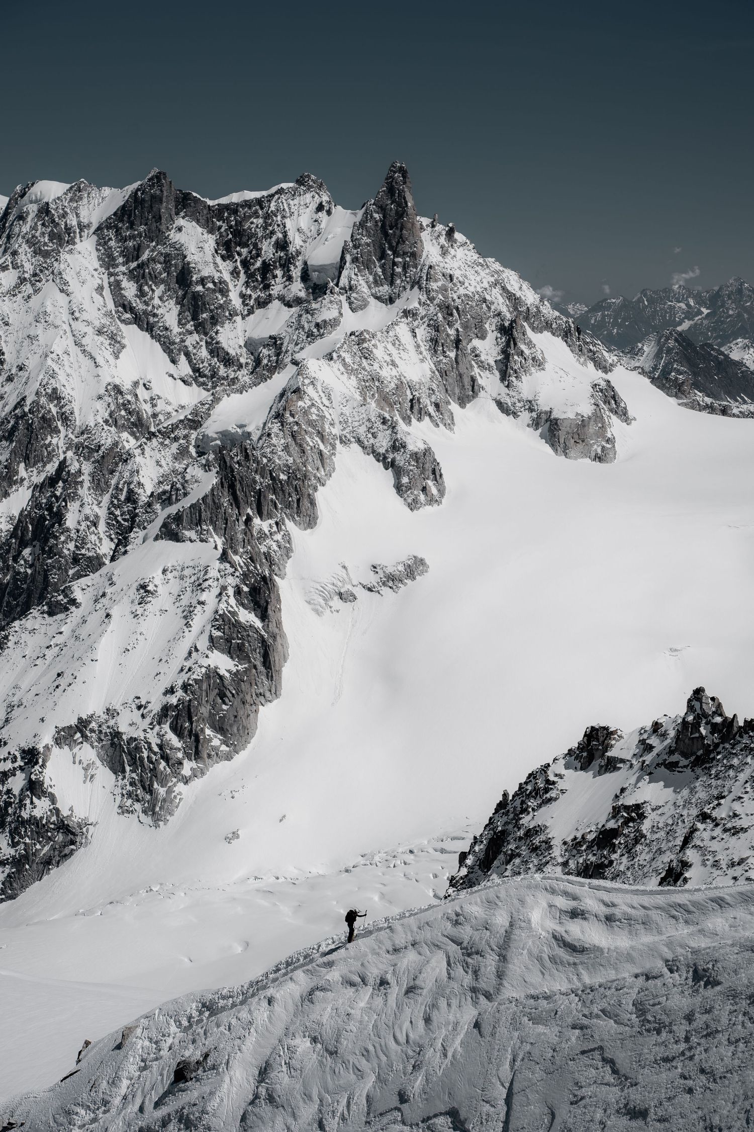 Lone mountaineer descends vast snow glacier below dramatic rocky snow-covered peaks in black and white.