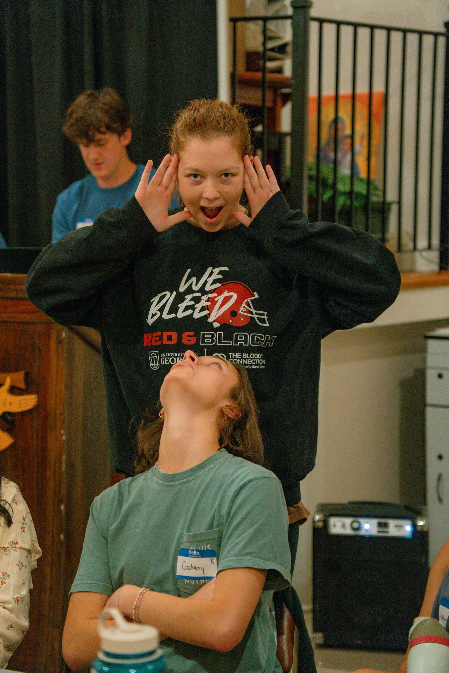 Young woman making a funny face with hands raised behind another girl at an indoor Catholic event in Athens, GA.