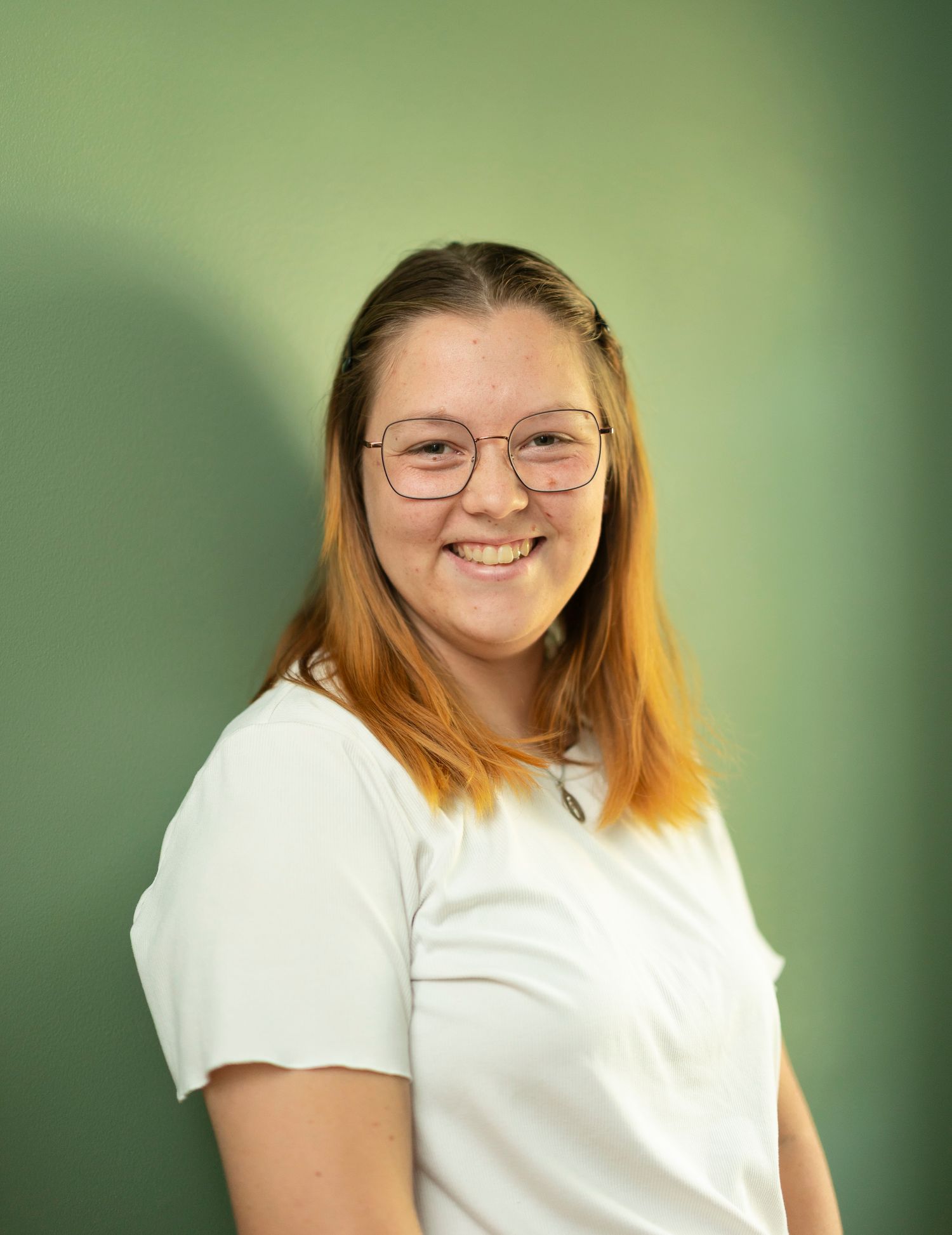 Smiling young woman with glasses and auburn hair wearing a white t-shirt against a green background.