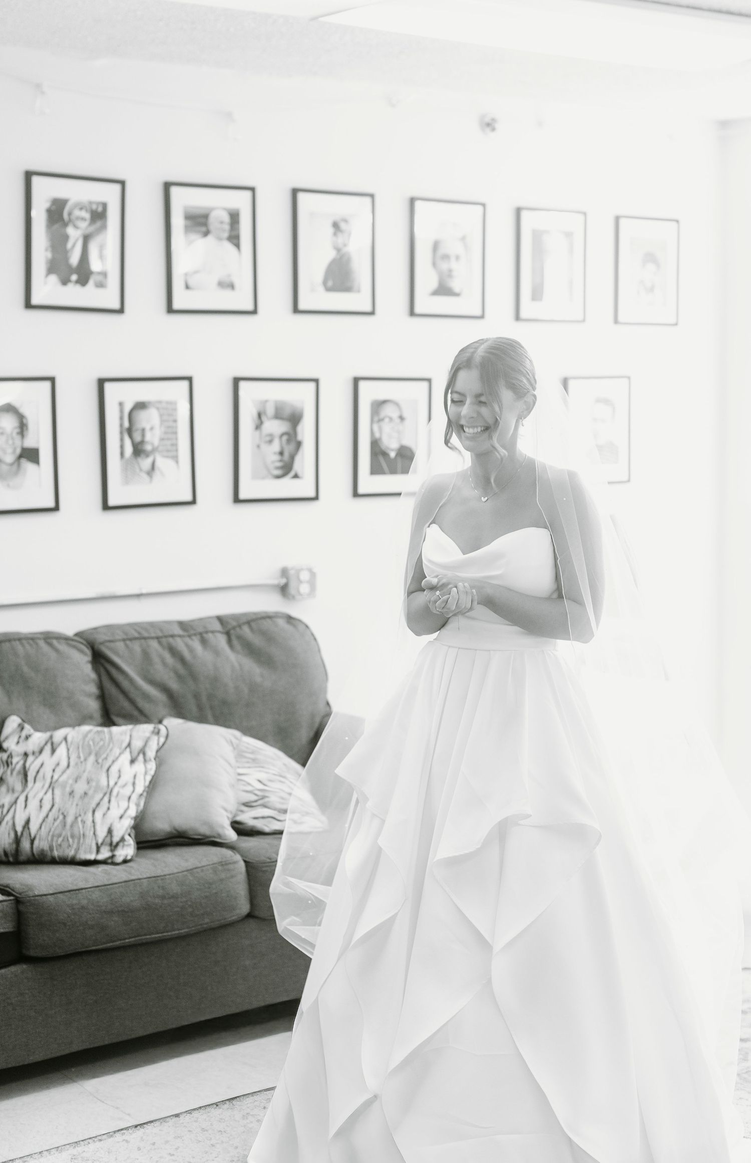 Smiling bride in a strapless ruffled white wedding gown stands in a room with framed photos on the wall before catholic wedding in Georgia.