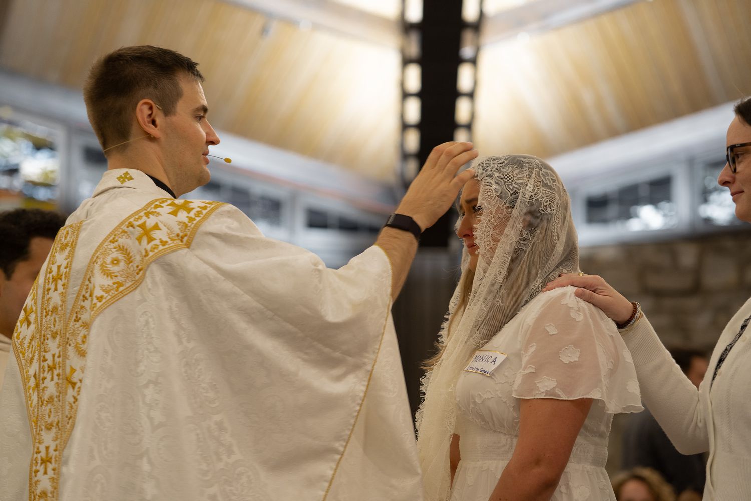 A young priest in gold vestments anoints a woman in white veil during Confirmation Mass at UGA's Catholic Center.