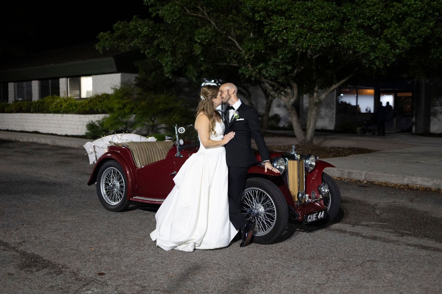 Bride and groom kissing beside a vintage red classic car on a street at night after their catholic wedding celebration.