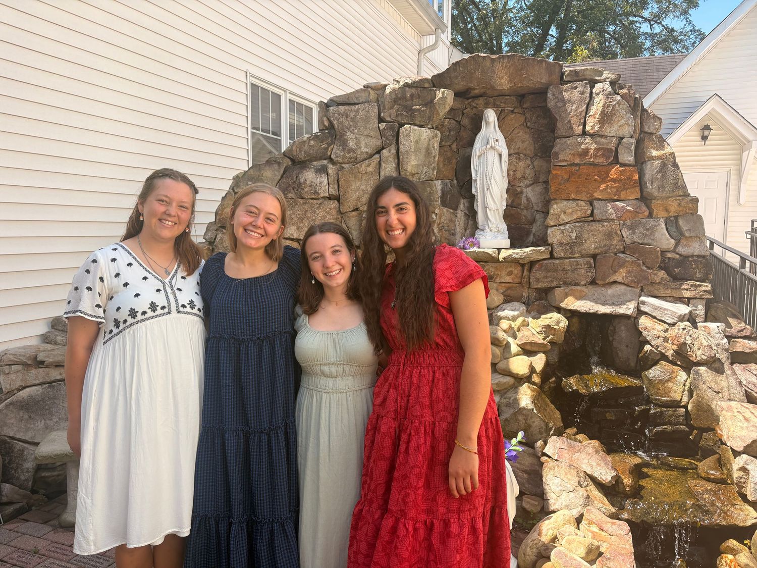 Four young women smiling together outdoors near a stone grotto with a Virgin Mary statue.