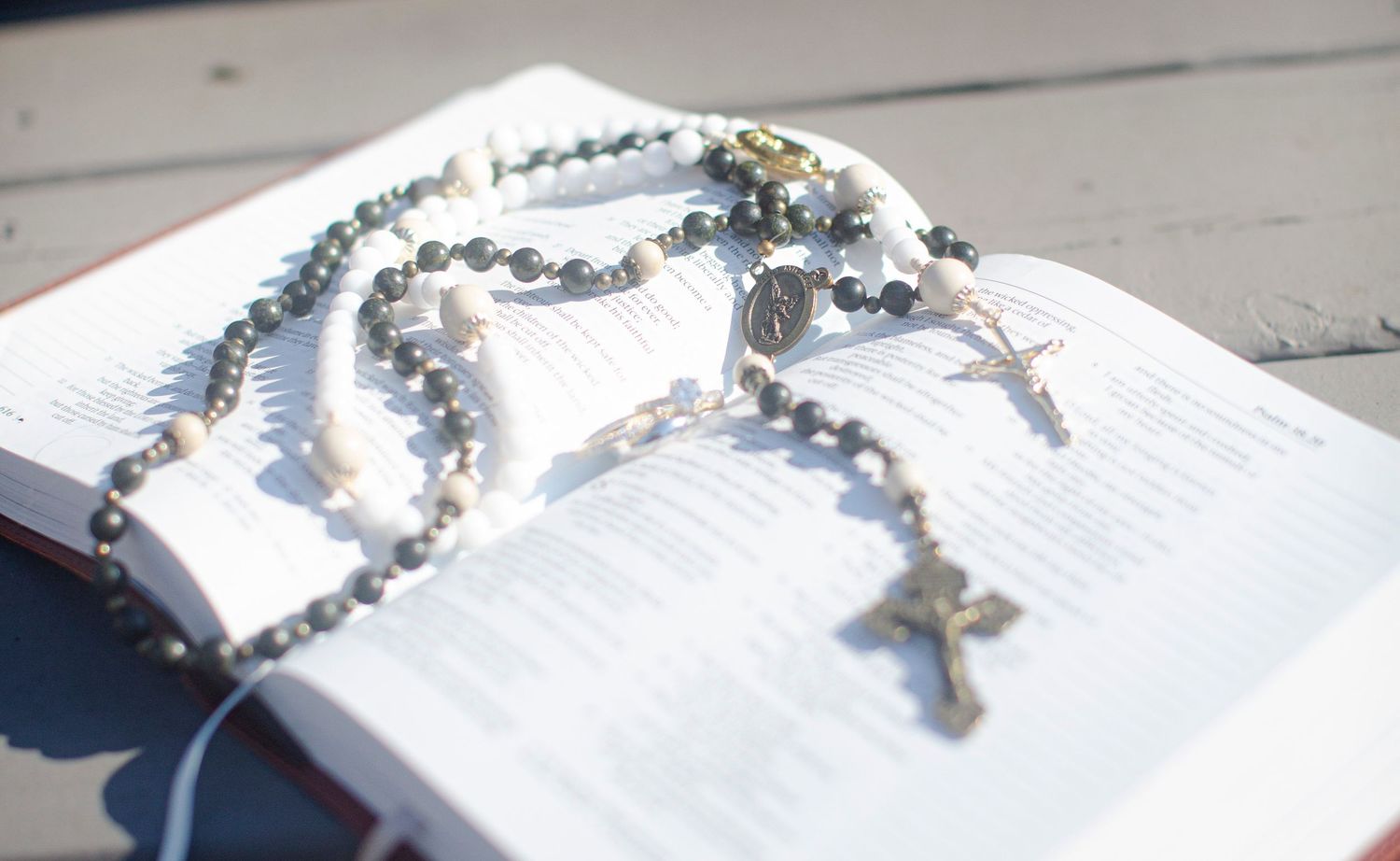Rosary beads with crucifix resting on open Bible pages, photographed outdoors in natural light.