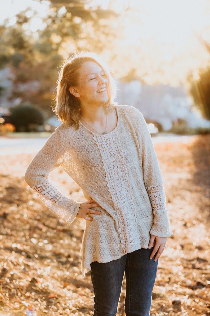Woman in beige knit sweater and jeans smiling outdoors in warm golden sunlight among fallen autumn leaves.