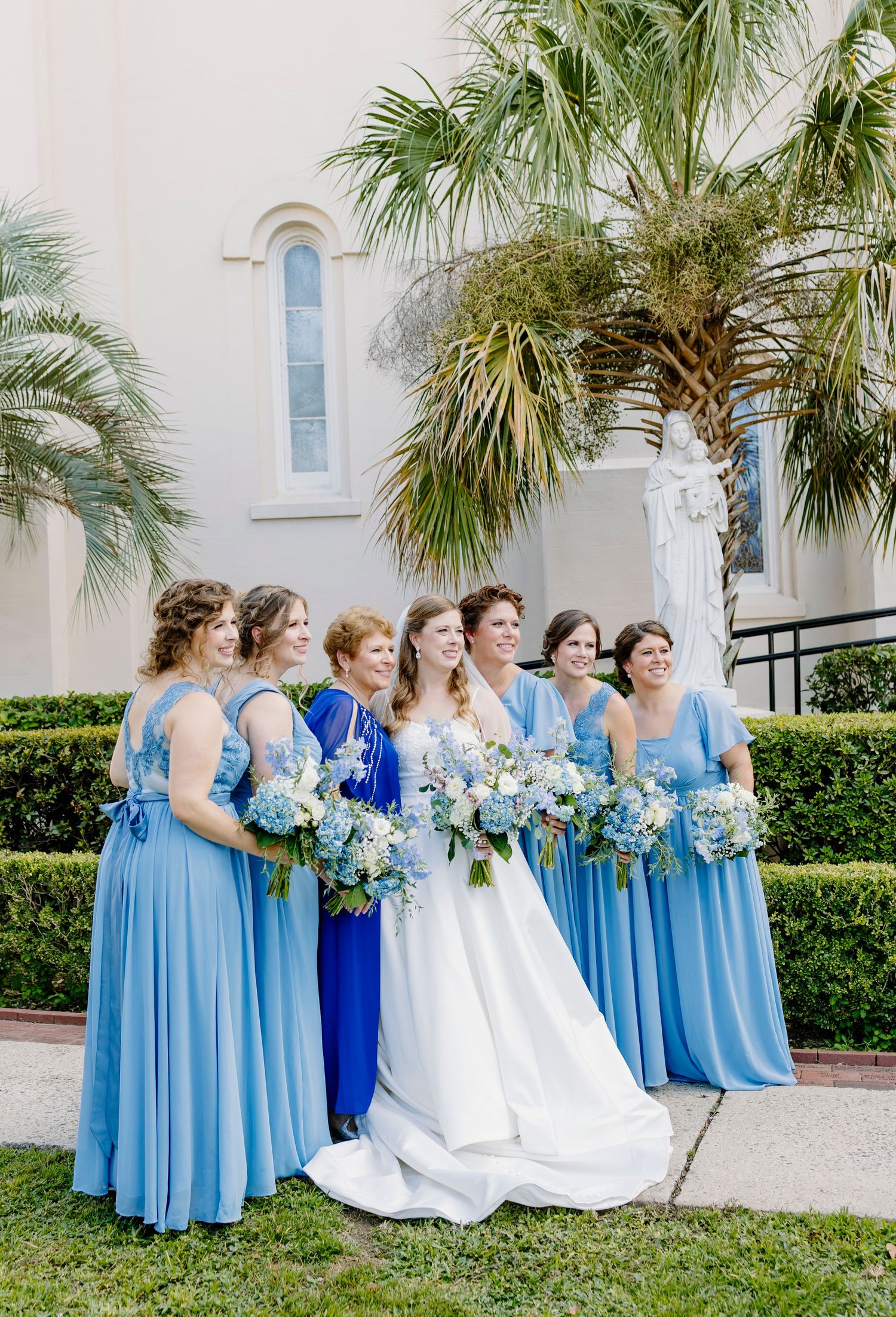 Bride in white gown poses with bridesmaids in light blue dresses holding floral bouquets outside a catholic church.