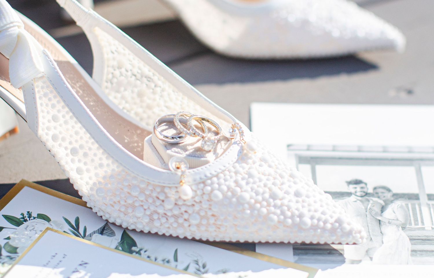 White pearl-covered bridal heels with gold wedding rings resting on top, displayed on a elegant wedding flat lay.