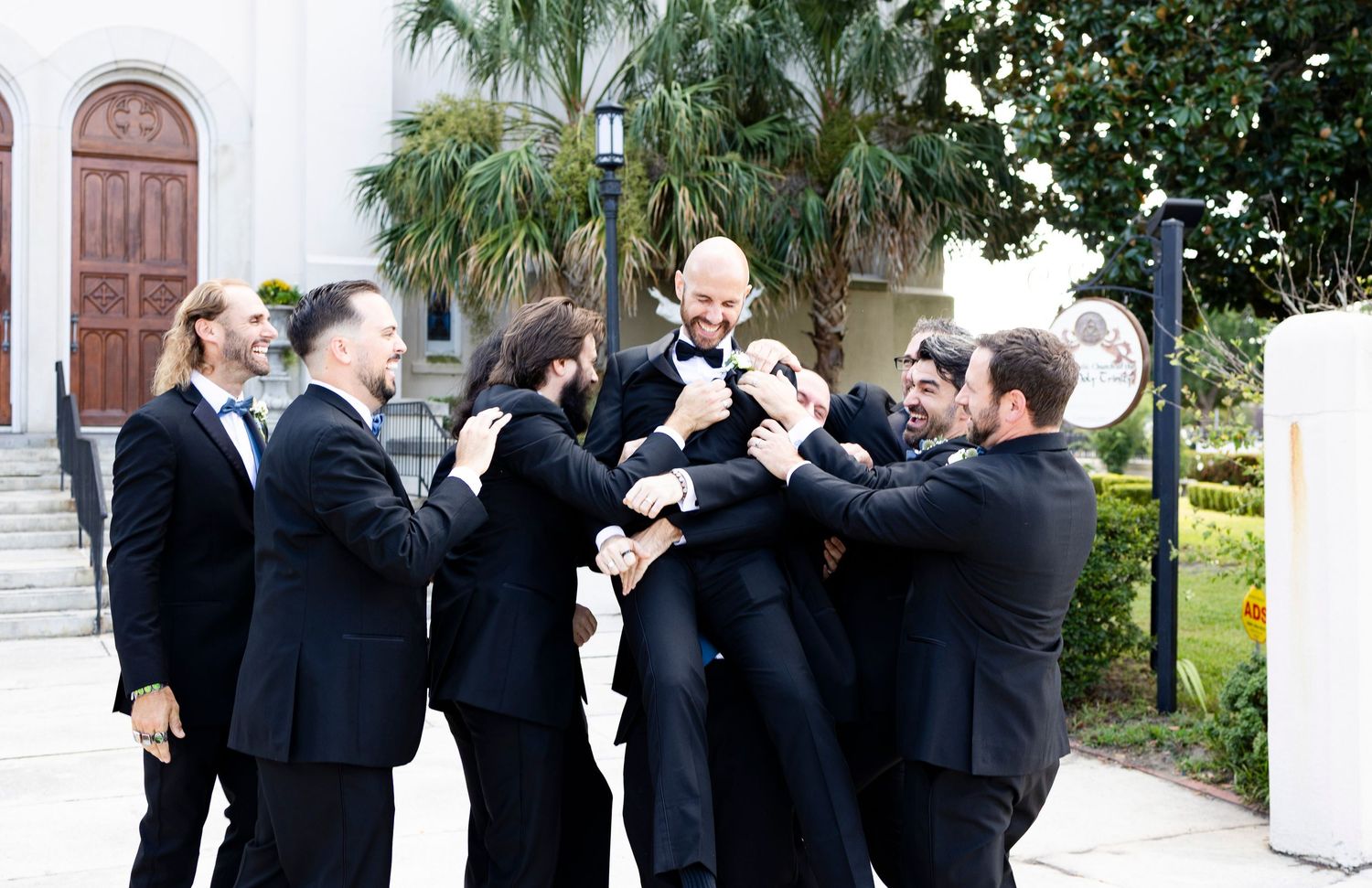 Groom surrounded by groomsmen in black tuxedos celebrating outdoors near a catholic church with palm trees in Augusta, GA.