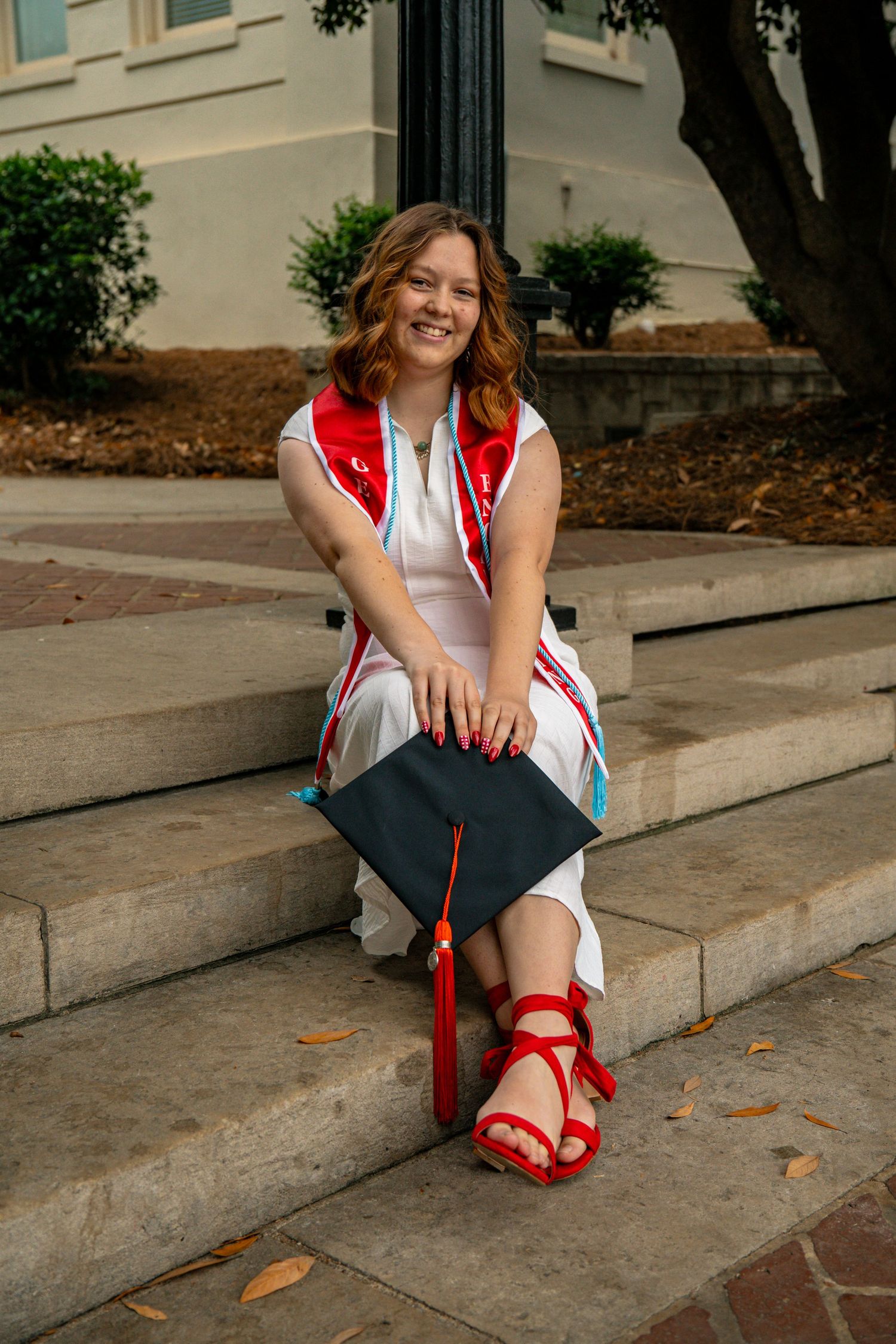 Smiling Catholic graduate in red sash sitting on steps holding black graduation cap in Athens, GA.