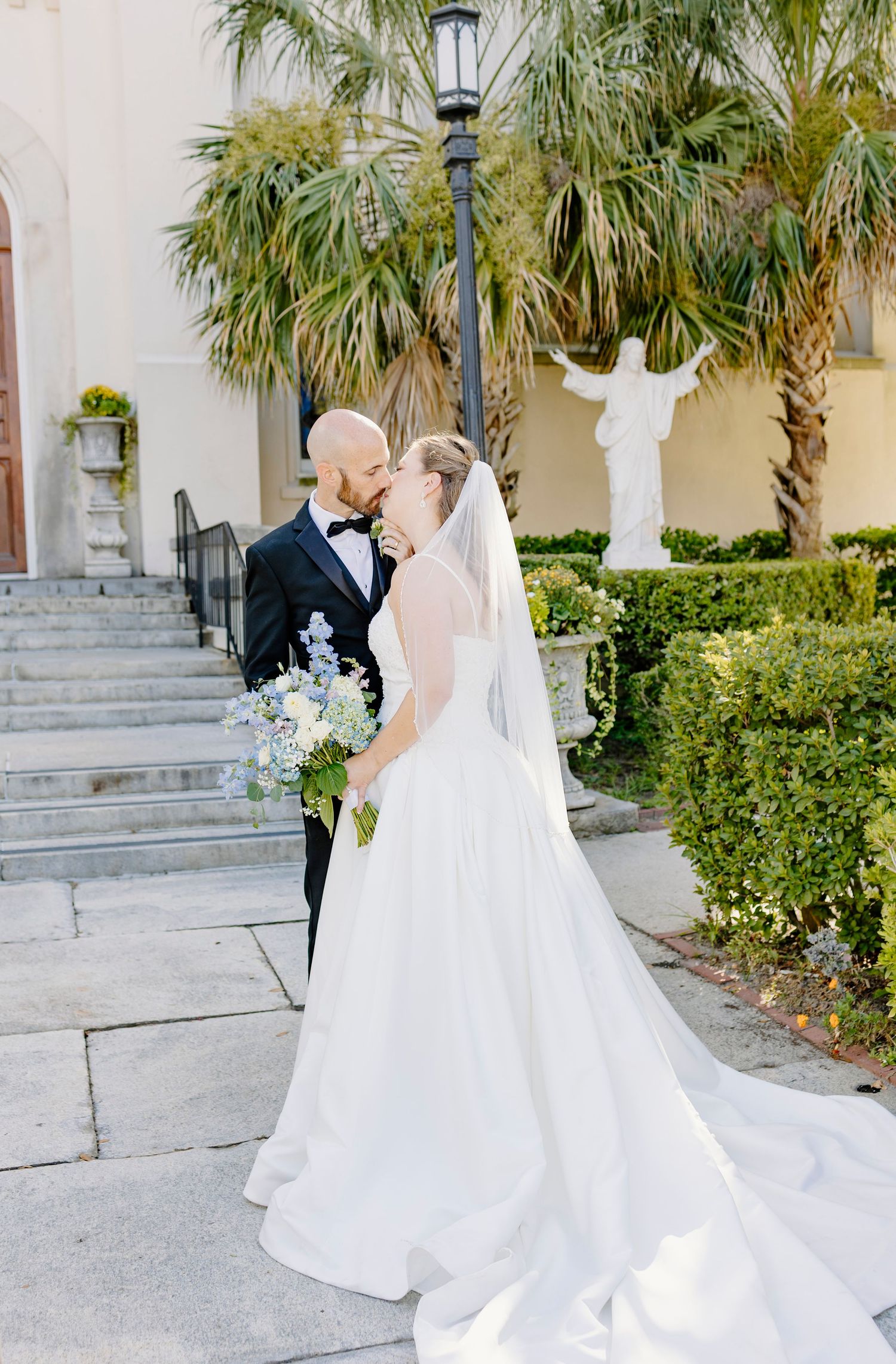 Bride and groom share a kiss outside a catholic church, with palm trees and a white statue in the background in Augusta, GA.