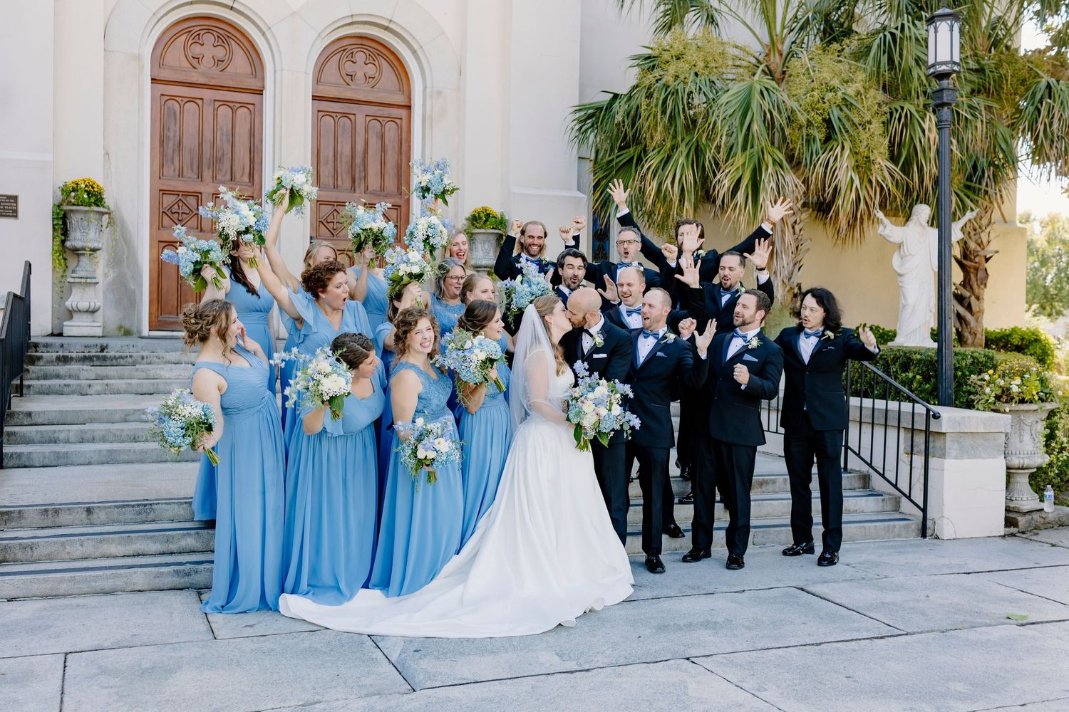 Wedding party celebrates outside a catholic church, bridesmaids in blue holding bouquets, groomsmen in black suits, bride in white gown.