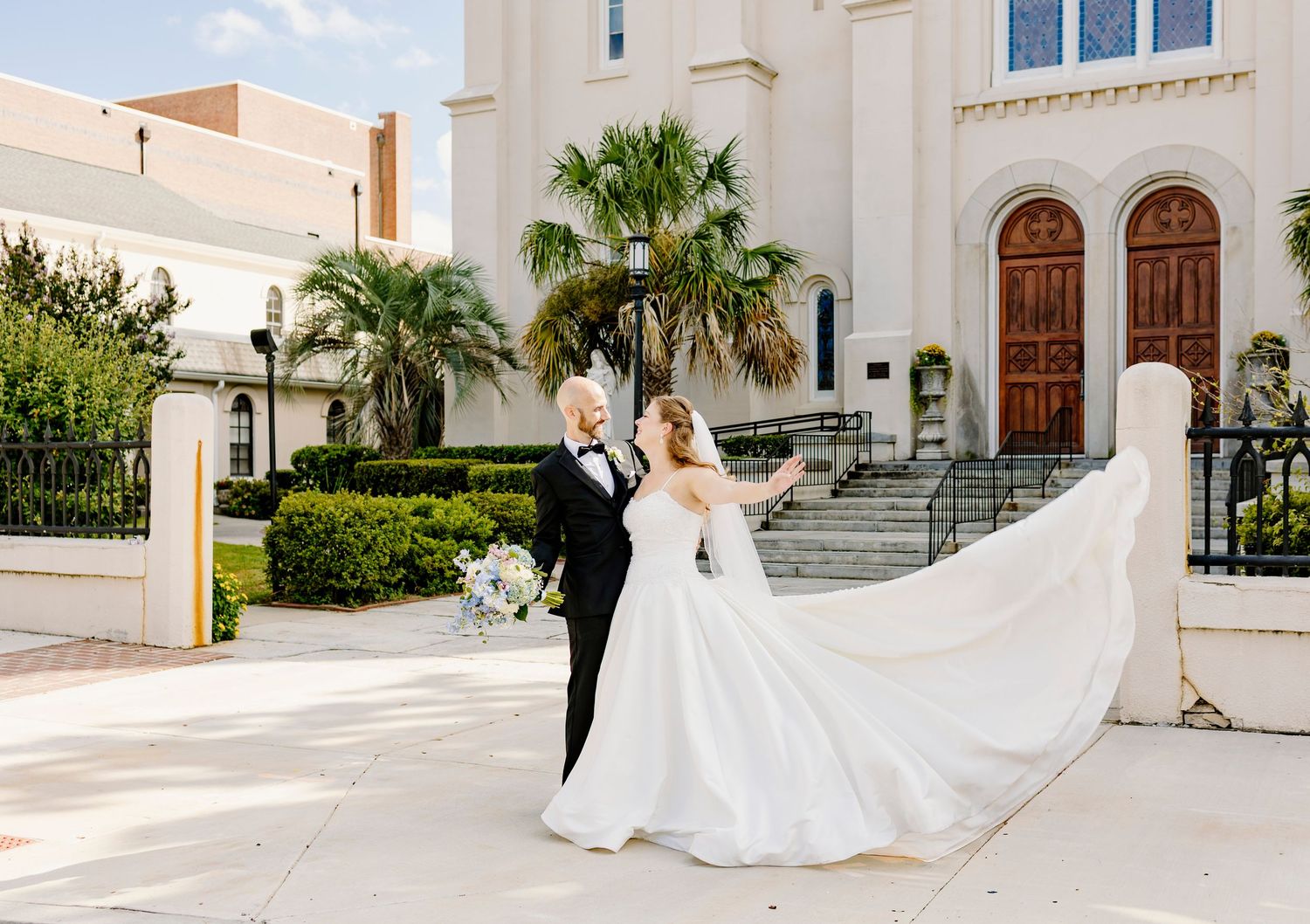 Bride in flowing white gown with veil blowing in wind poses with groom in black tux outside elegant white catholic church in Augusta, GA.