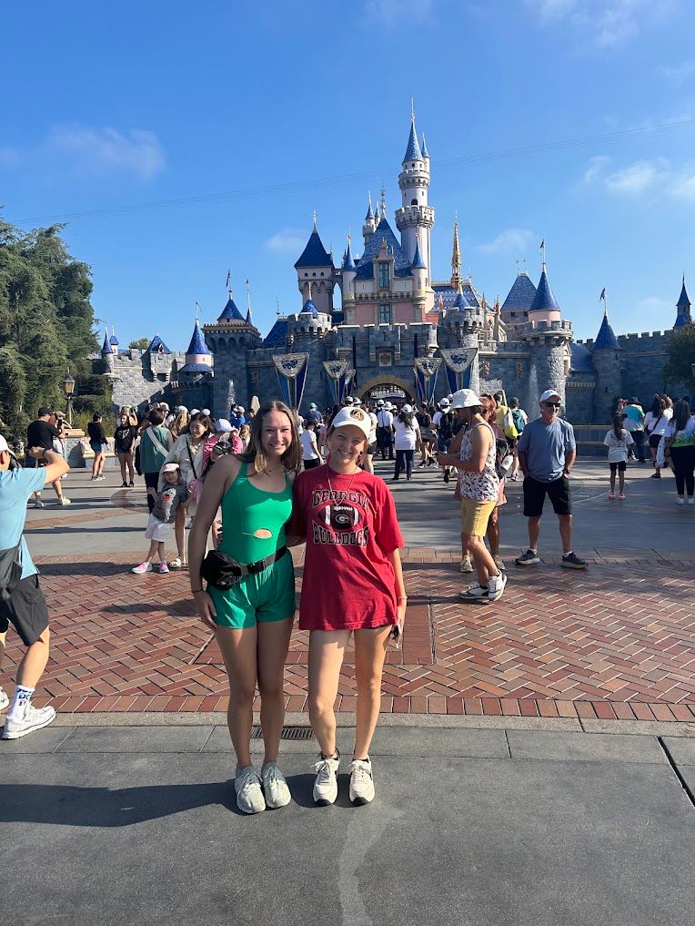 Two girls smiling in front of Sleeping Beauty Castle at Disneyland on a bright sunny day.