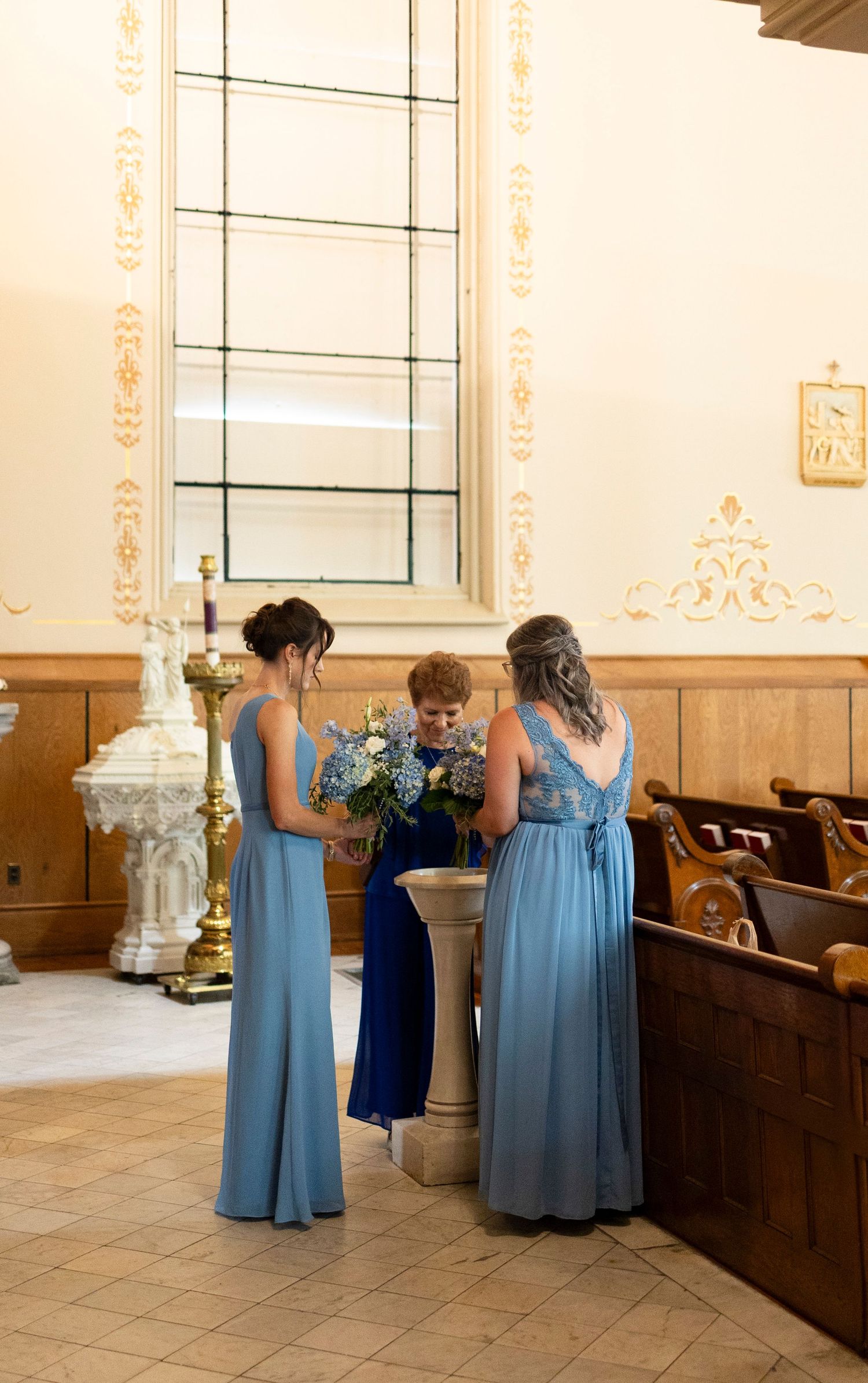 Two bridesmaids in blue gowns stand with a guest near a baptismal font inside an elegant catholic church in Augusta, GA.