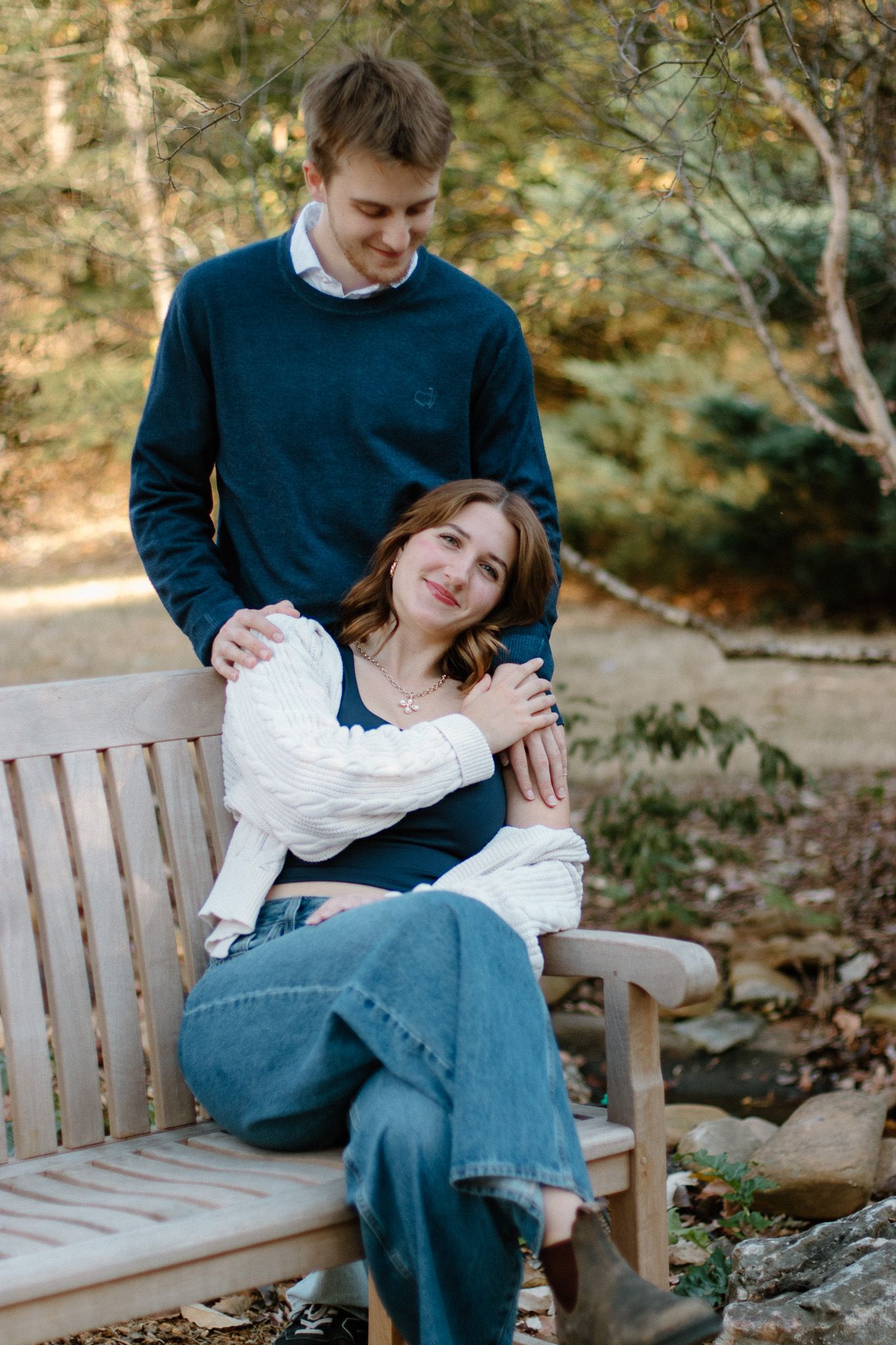 A young catholic couple poses on a wooden bench in a park, the man standing behind the seated woman in casual attire in Athens, GA.
