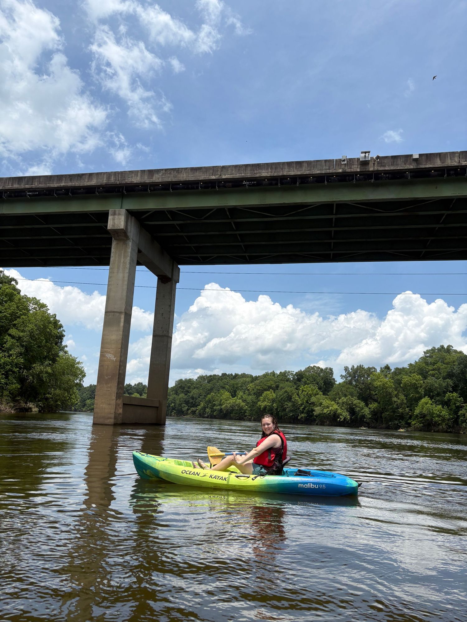 Woman kayaking on a calm river under a large bridge on a partly cloudy day.