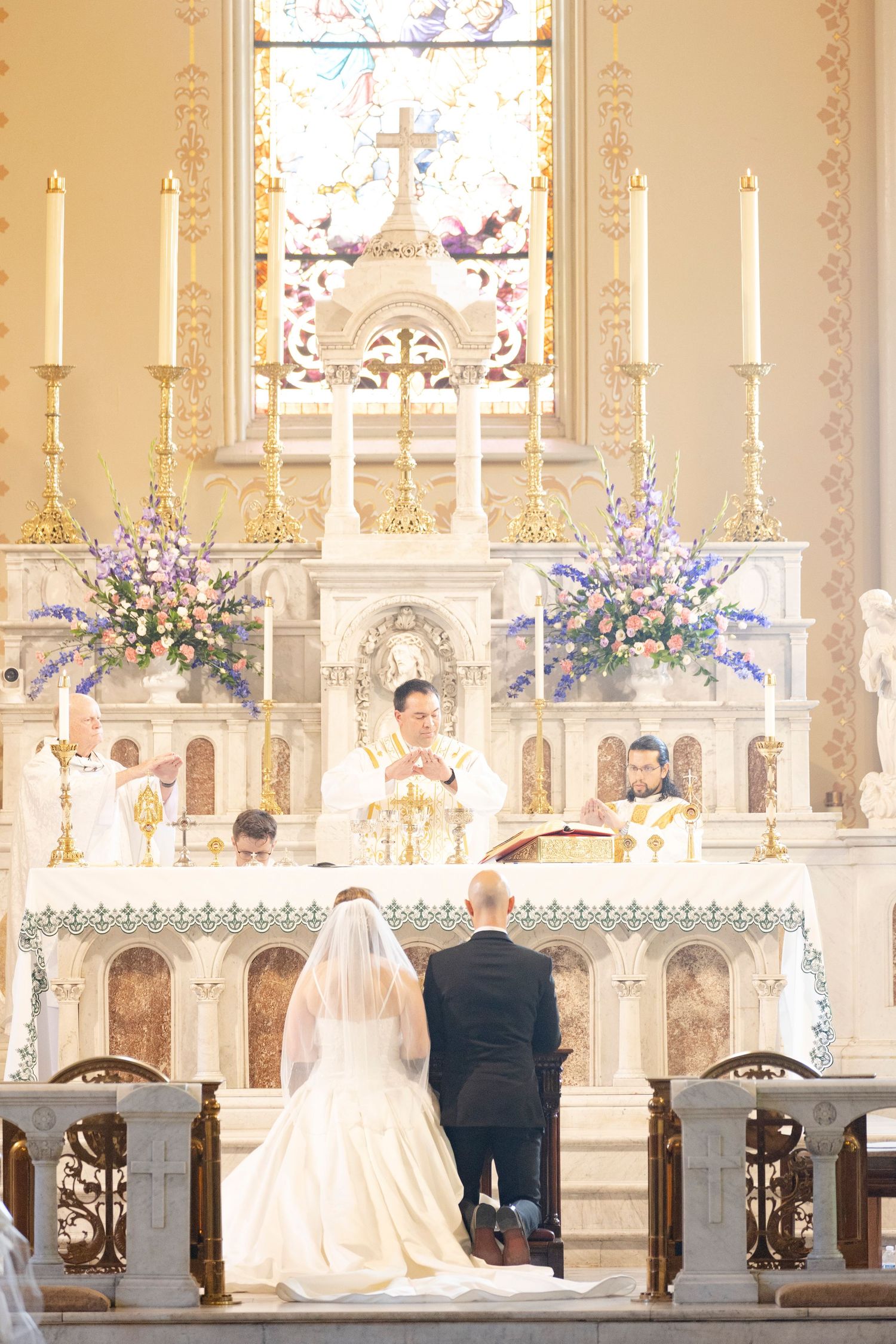 Bride and groom kneel at ornate Catholic church altar during wedding ceremony with priest and floral arrangements in Augusta, GA.