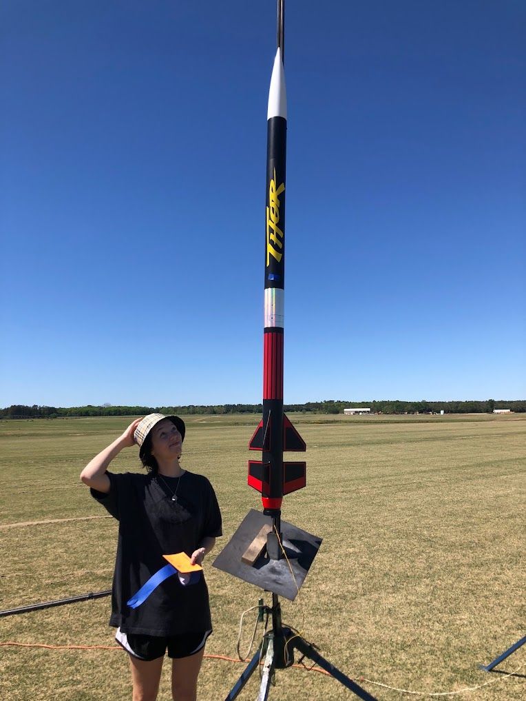 Young woman standing beside a tall model rocket on a launch pad in an open field.