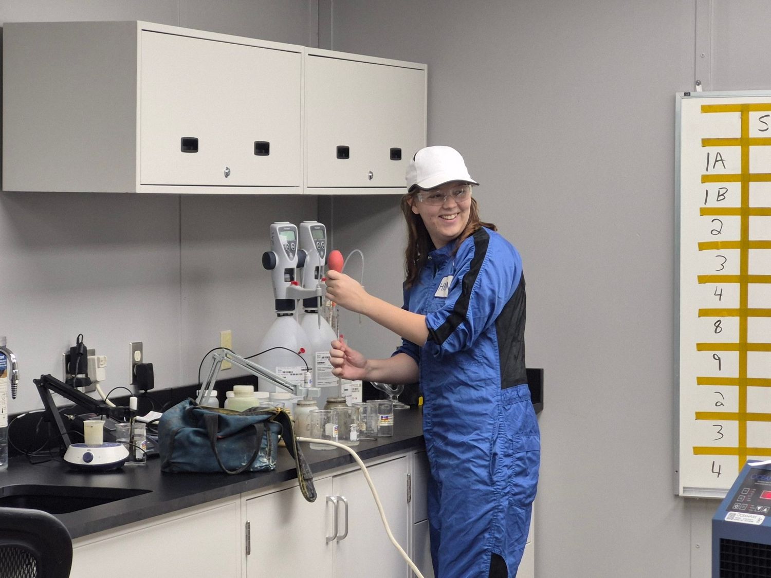Young woman in blue lab coat and hard hat working with equipment in a laboratory.