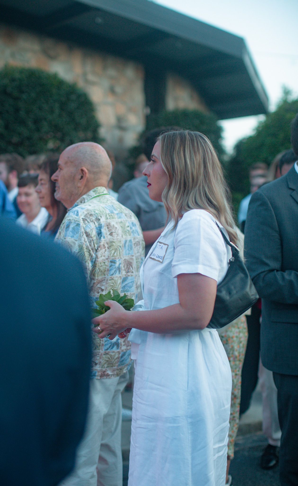 A blonde woman in a white dress with a black purse stands among a crowd at a Catholic Easter Vigil Mass in Athens, GA