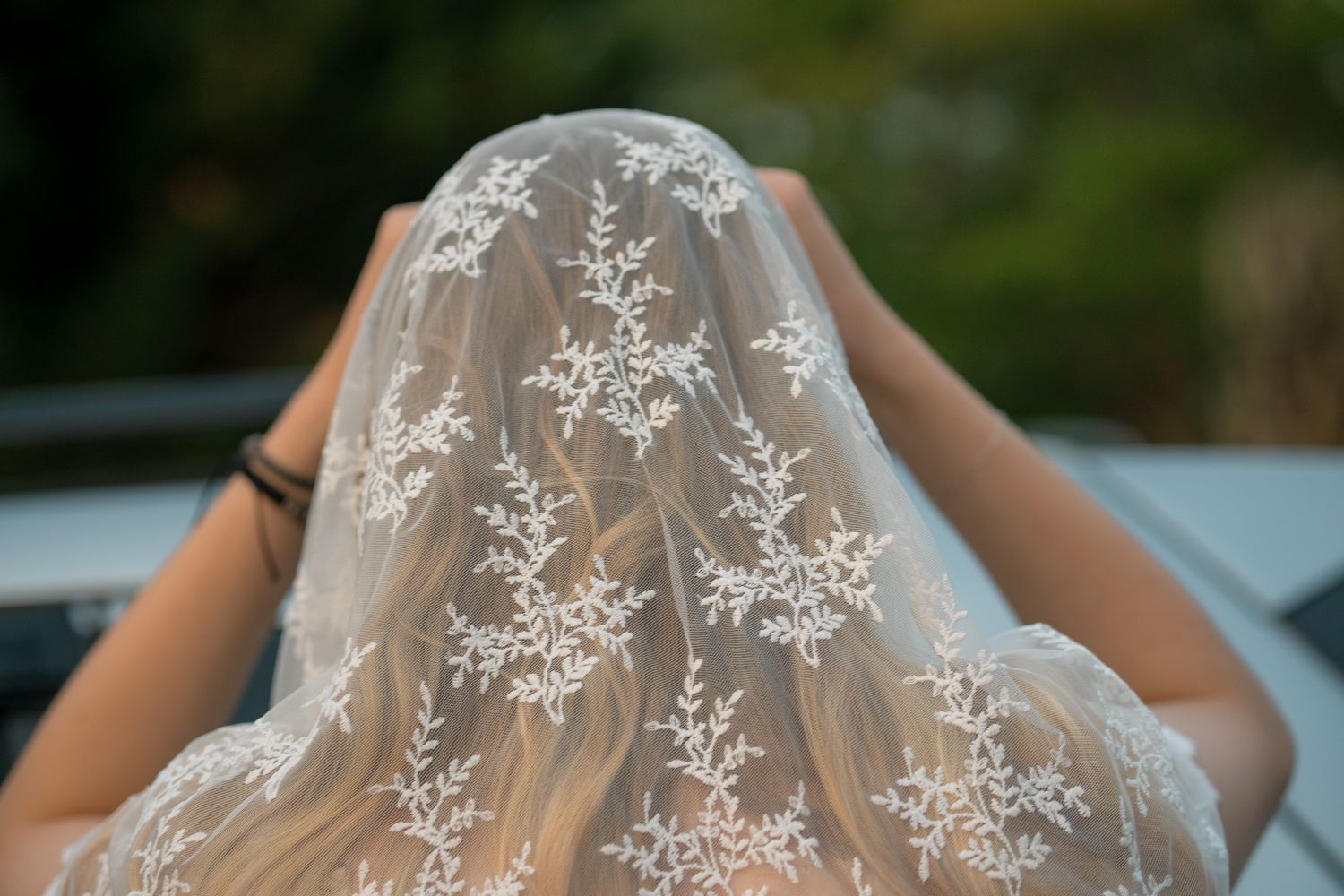 Bride wearing an elegant lace wedding veil with floral embroidery over long blonde hair outdoors.