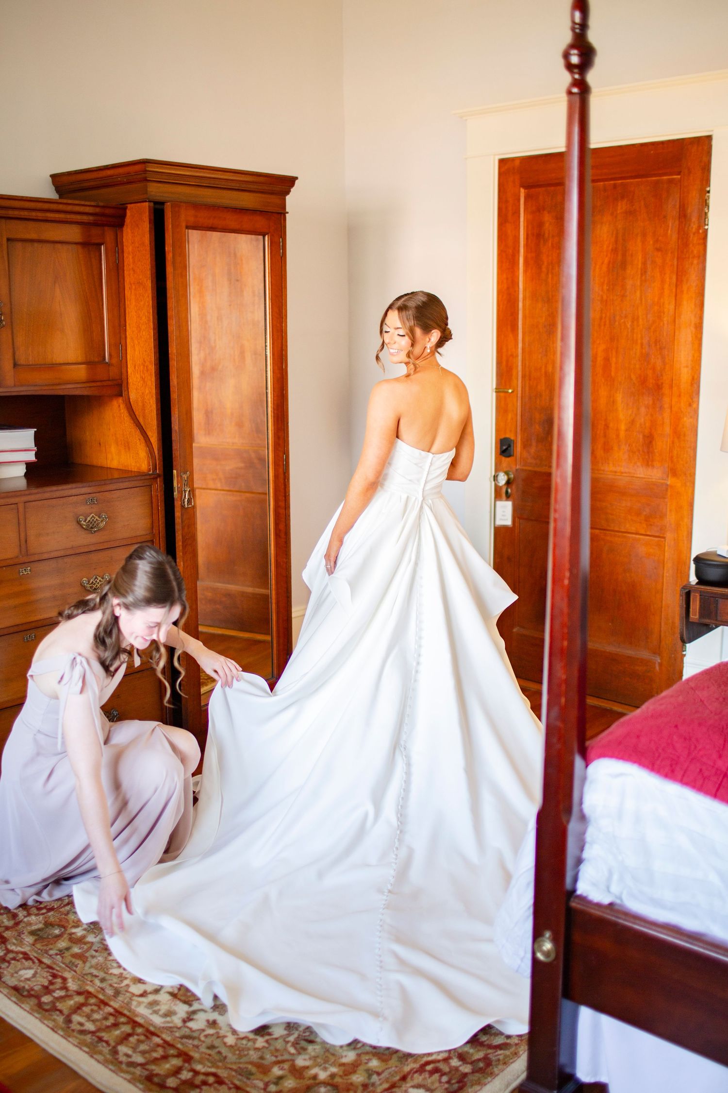 Bridesmaid adjusting the train of a catholic bride's white wedding gown in an elegant bedroom with wood furniture.
