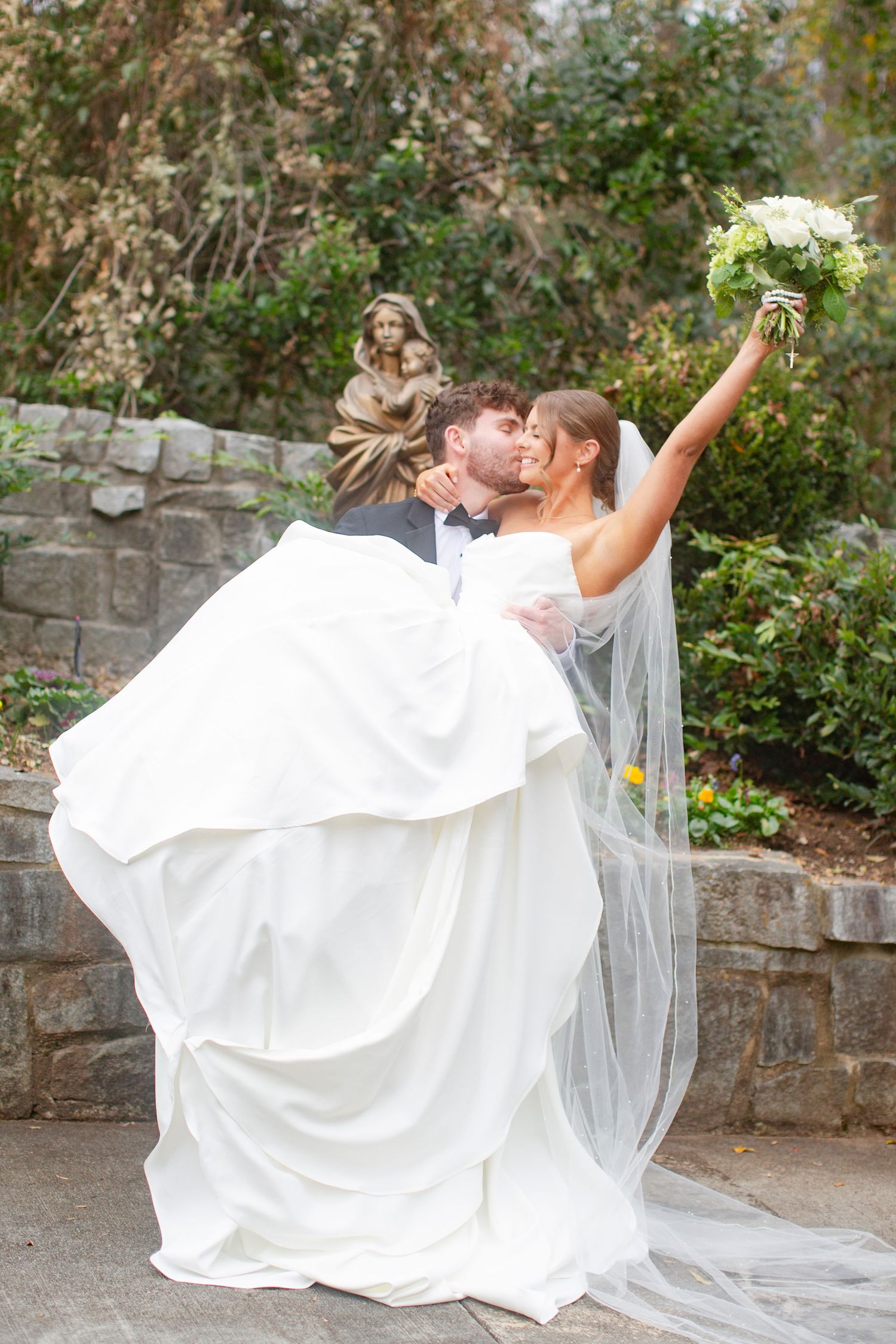 Groom lifts bride in white gown as they kiss in a garden, bride raising bouquet, stone statue behind them after catholic wedding.