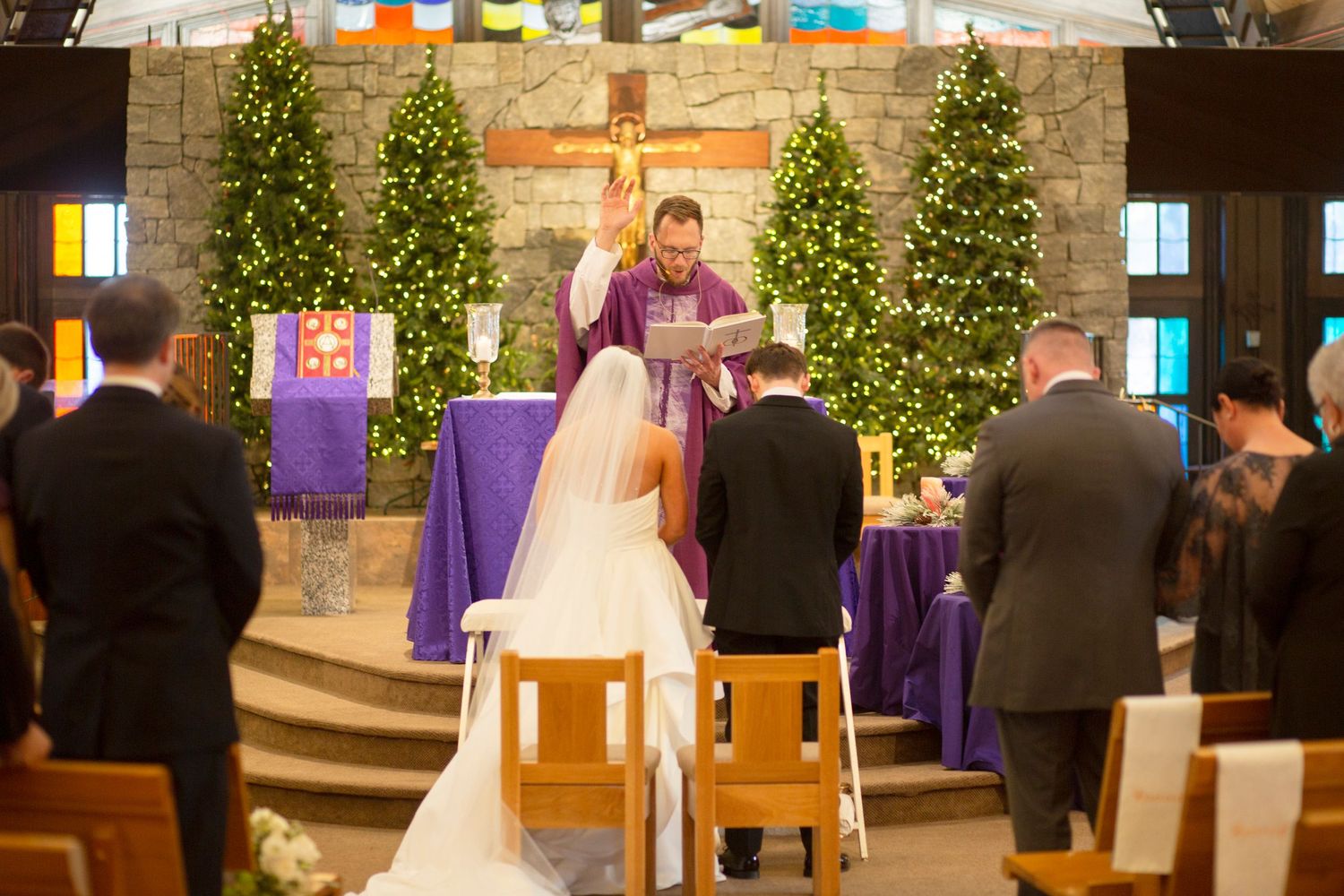 A couple stands before a priest at a decorated Catholic altar during a wedding ceremony with Christmas trees in Athens, GA.