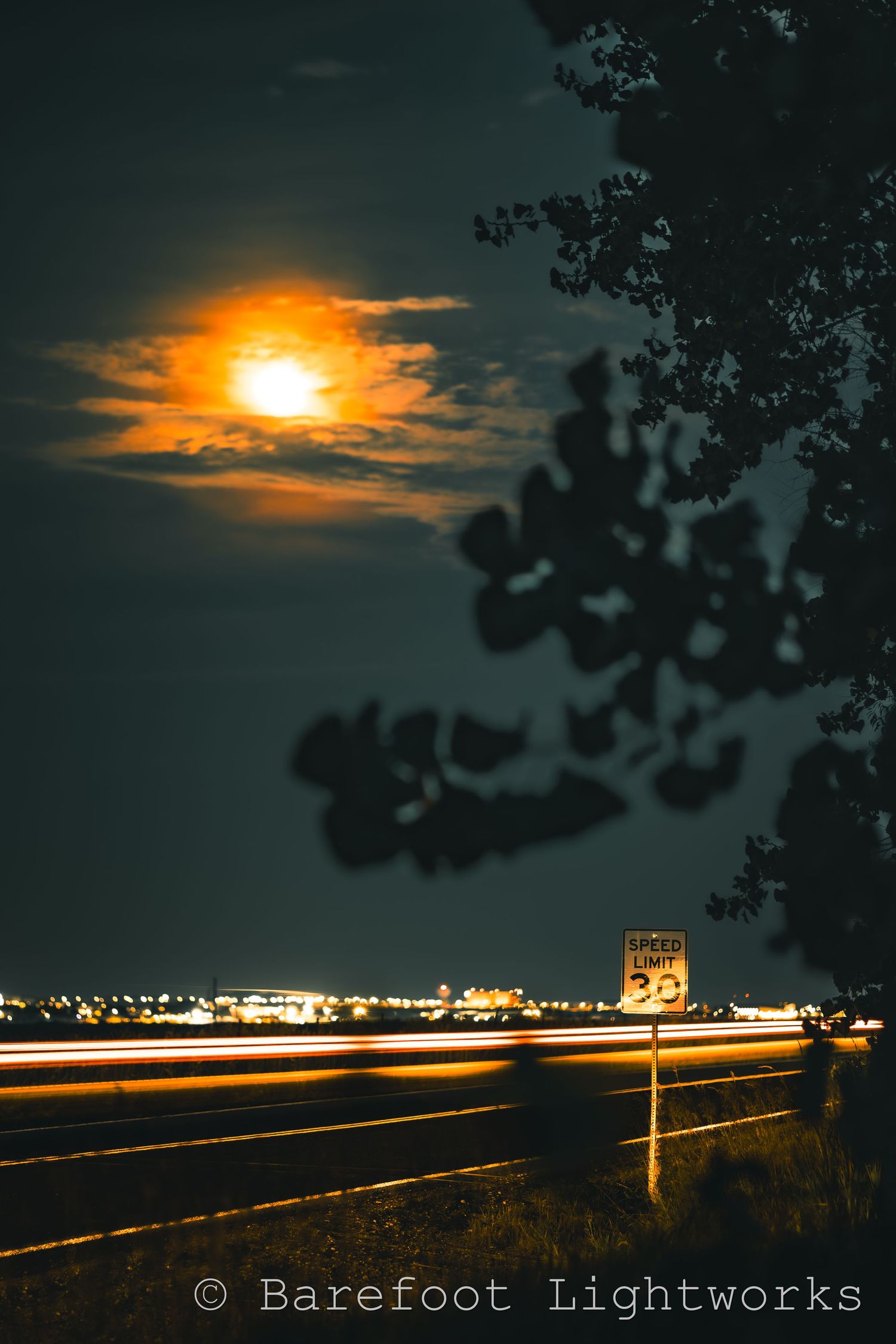 A photo of the moon and night sky near a road. A car leaves light streaks from it's lights as it drives by