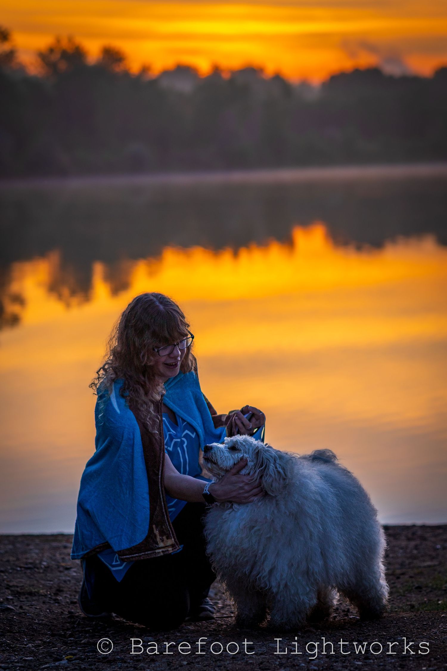 A photo of a woman with wavy blond hair and glasses with her dog in the morning sunrise near the lake.
