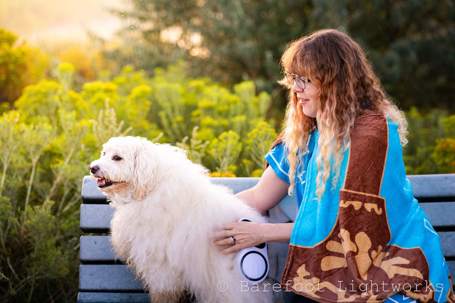 A photo of a woman with wavy blond hair and glasses with her dog in the morning sunlight.
