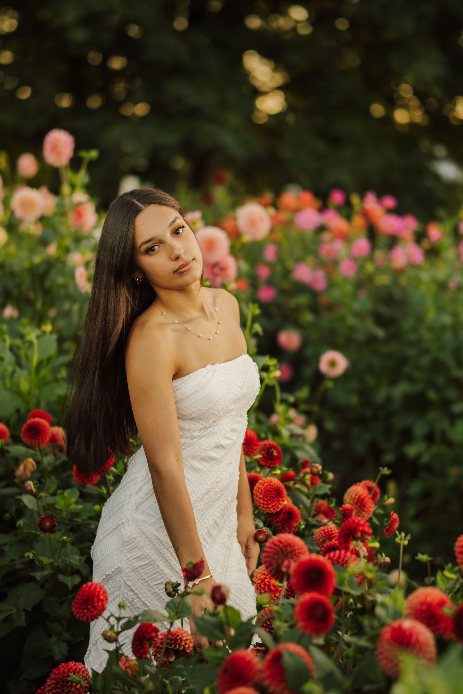 Young woman with long dark hair in a white strapless dress poses among blooming red and pink dahlias in a lush garden.