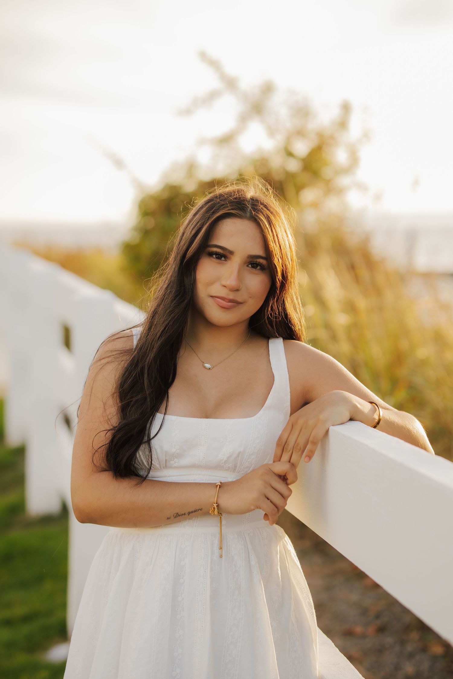 Young woman with long dark hair in a white sundress leans on a white wooden fence at golden hour, with tall grass behind her.
