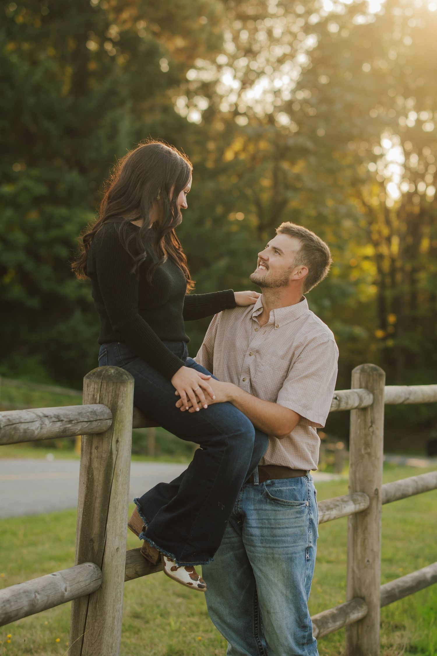 Couple smiling at each other as a woman sits on a wooden fence and a man stands beside her in golden evening light.